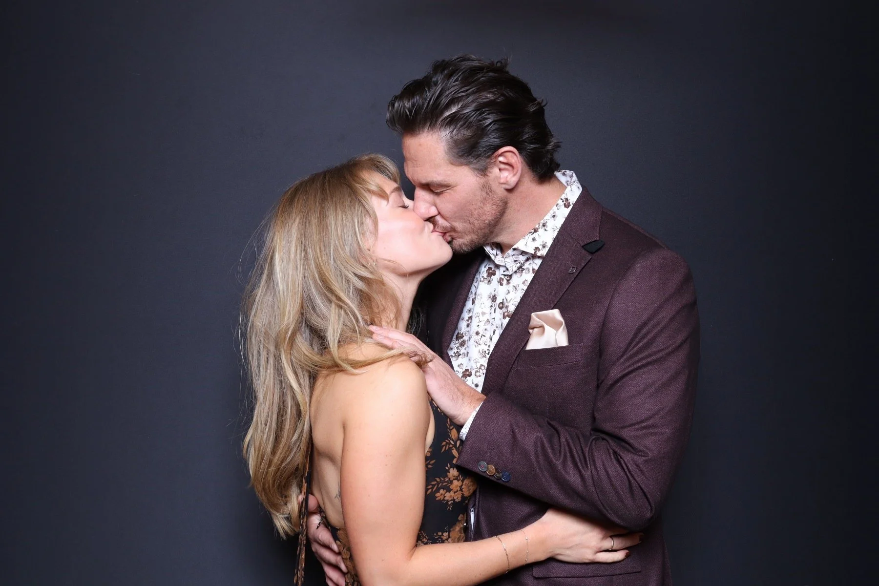 A man and woman are kissing, with the man's hand on the woman's neck and the woman's hand on the man's waist, against a dark background.