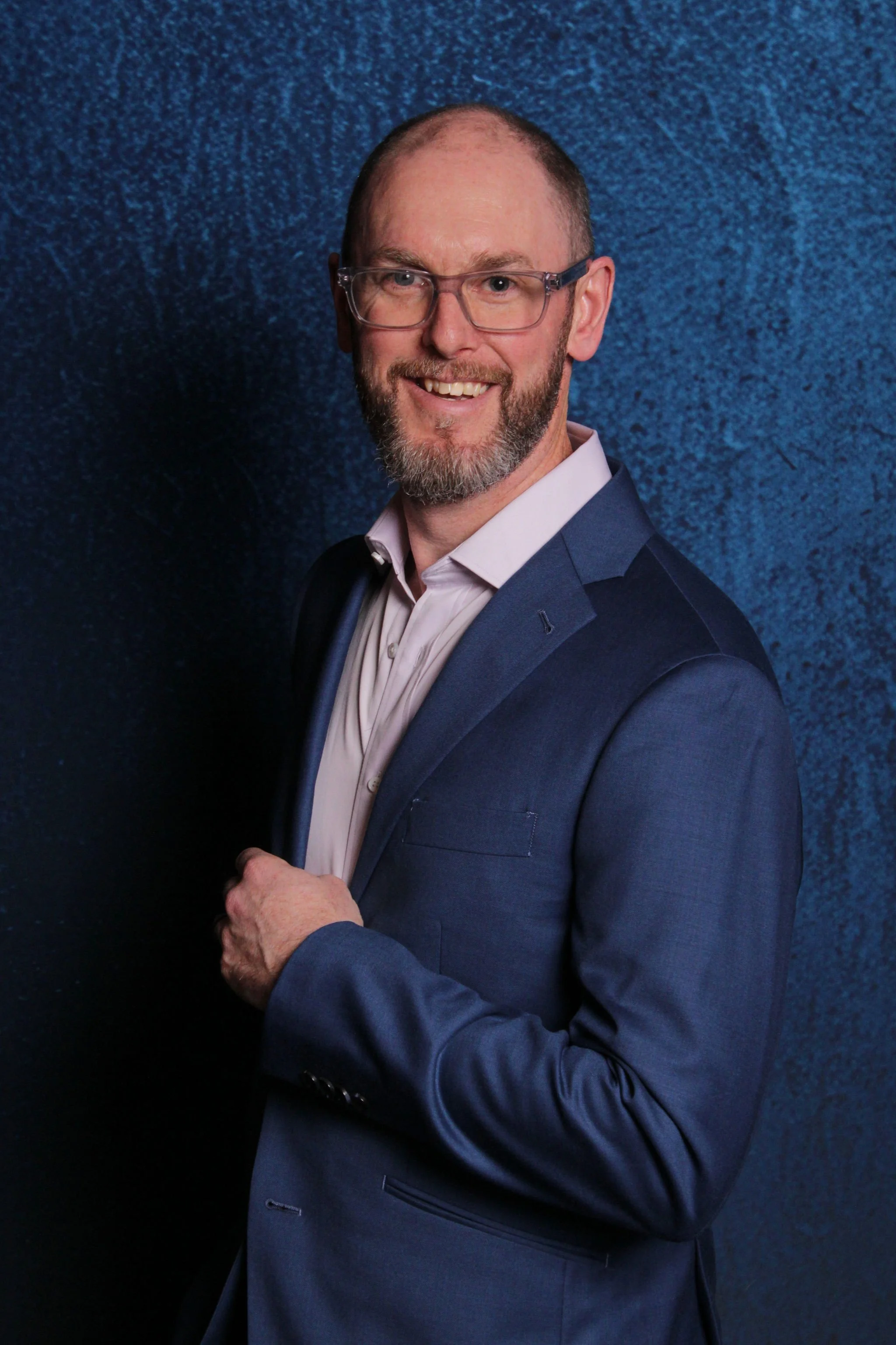 A middle-aged man with glasses, a beard, and short hair, dressed in a dark blue suit and white shirt, standing against a textured blue background.
