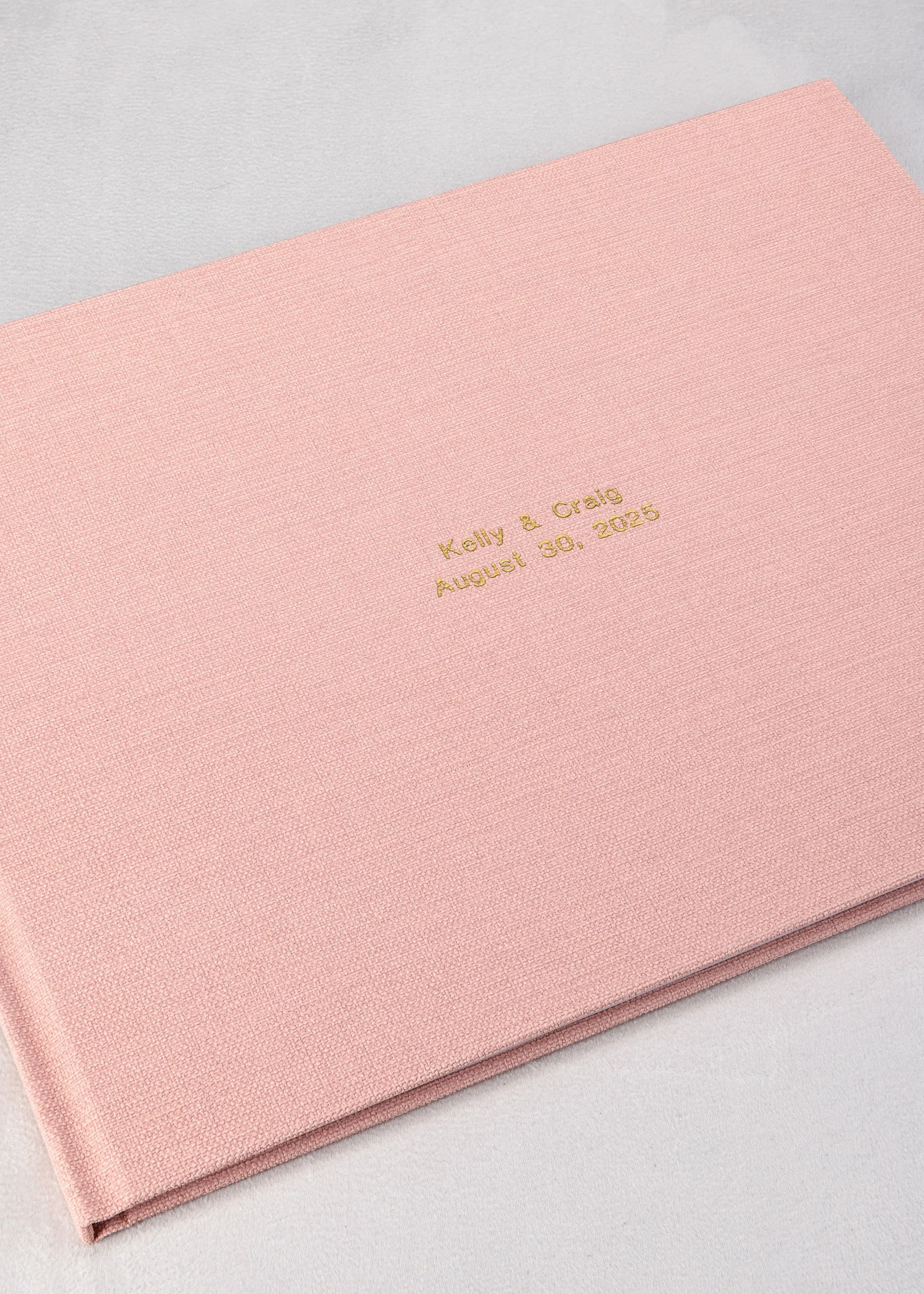 A pink textured wedding album with gold embossed text reading 'Kelly & Craig August 30, 2026' on the cover.