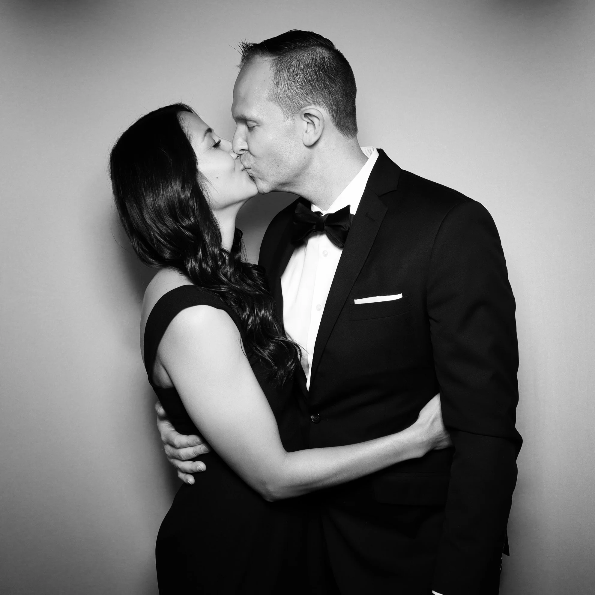 Black and white portrait of a couple in formal attire sharing a kiss, with the woman wearing a sleeveless dress and the man wearing a tuxedo.