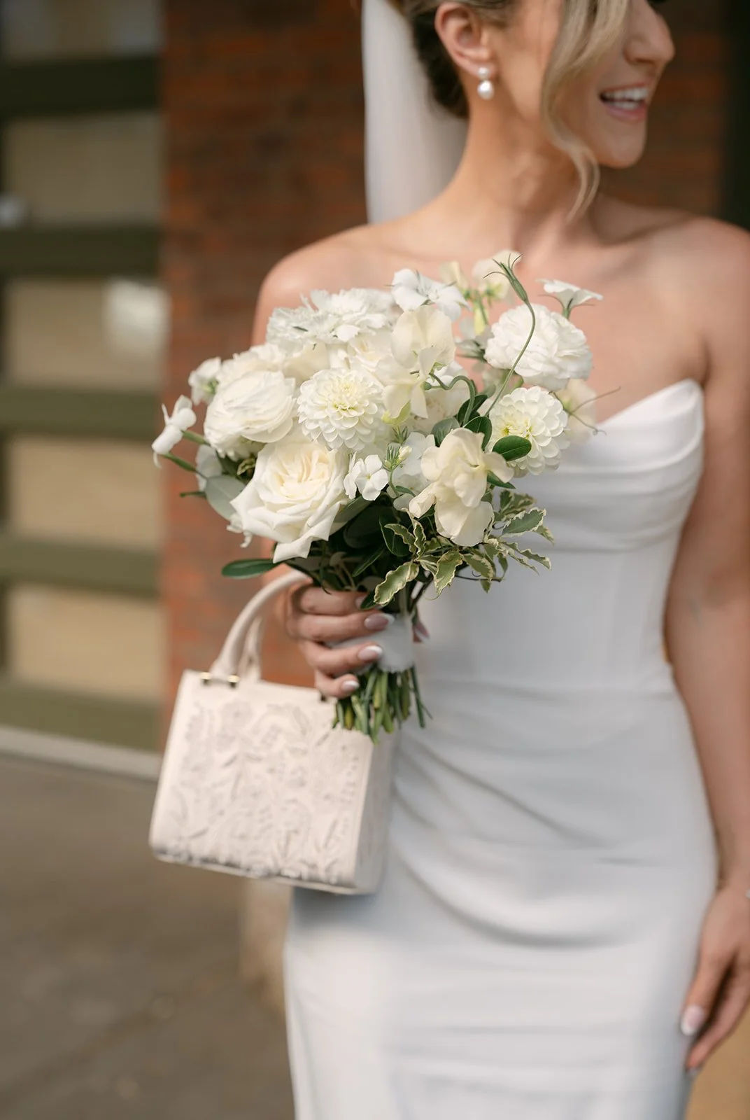 A woman in a white wedding dress holding a bouquet of white flowers and a small cream-colored purse with a floral pattern.