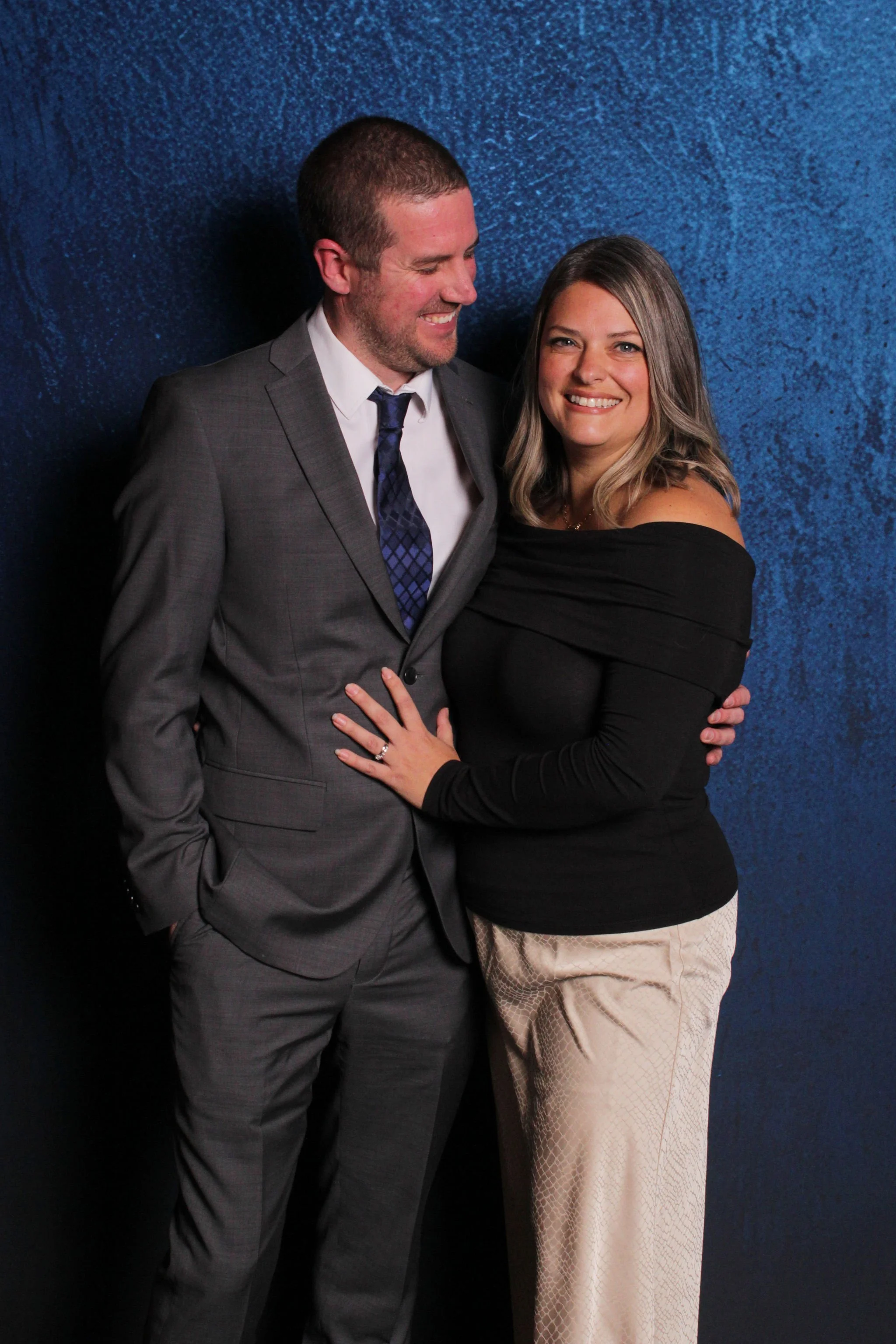 A man in a gray suit and a woman in a black off-shoulder top and cream-colored pants pose together against a blue textured background. They are smiling, with the woman’s hand on the man’s chest, showing an engagement ring.