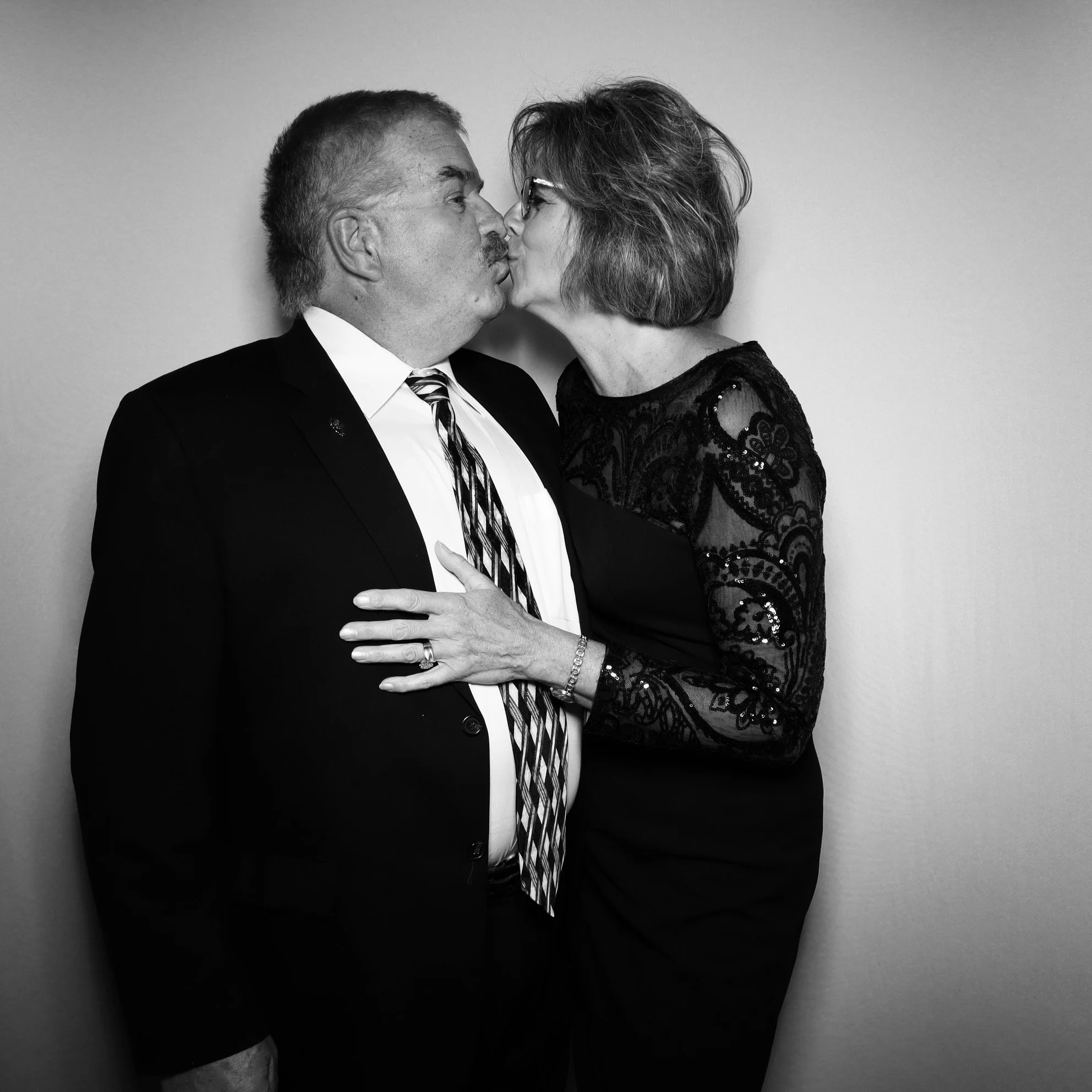 A black and white photo of an older man and woman kissing. The man is wearing a suit and tie, the woman is in a dress with lace sleeves and glasses.