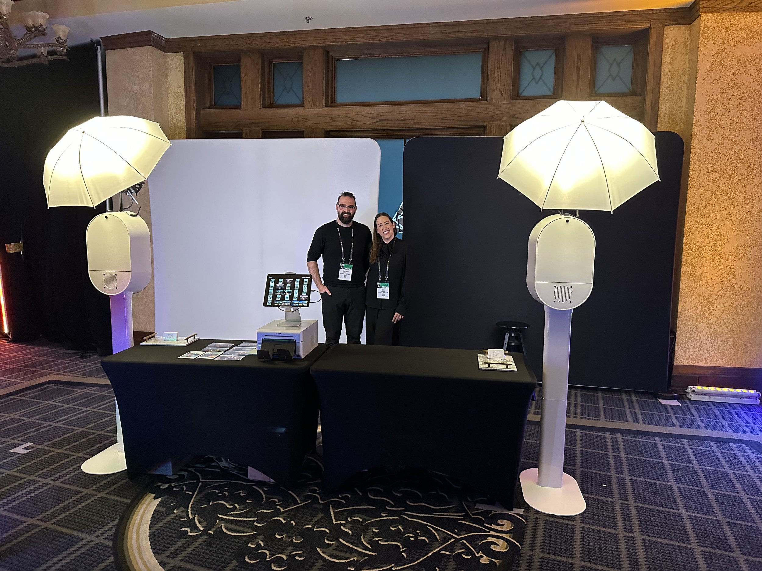 Two people, a man and a woman, standing together at a conference booth with photo props and lighting equipment
