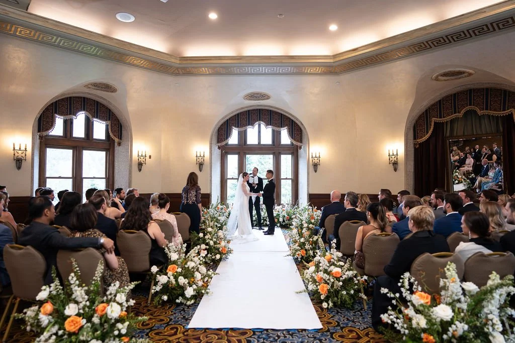 Bride and groom exchanging vows at a wedding ceremony in an elegant ballroom with floral arrangements and an aisle.