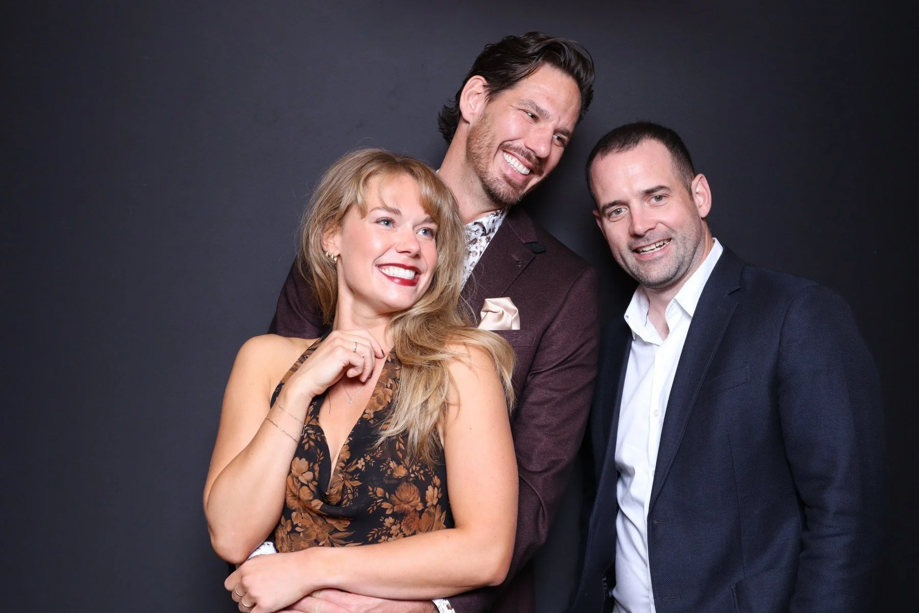 Three people smiling and posing together against a dark background, dressed in formal attire.