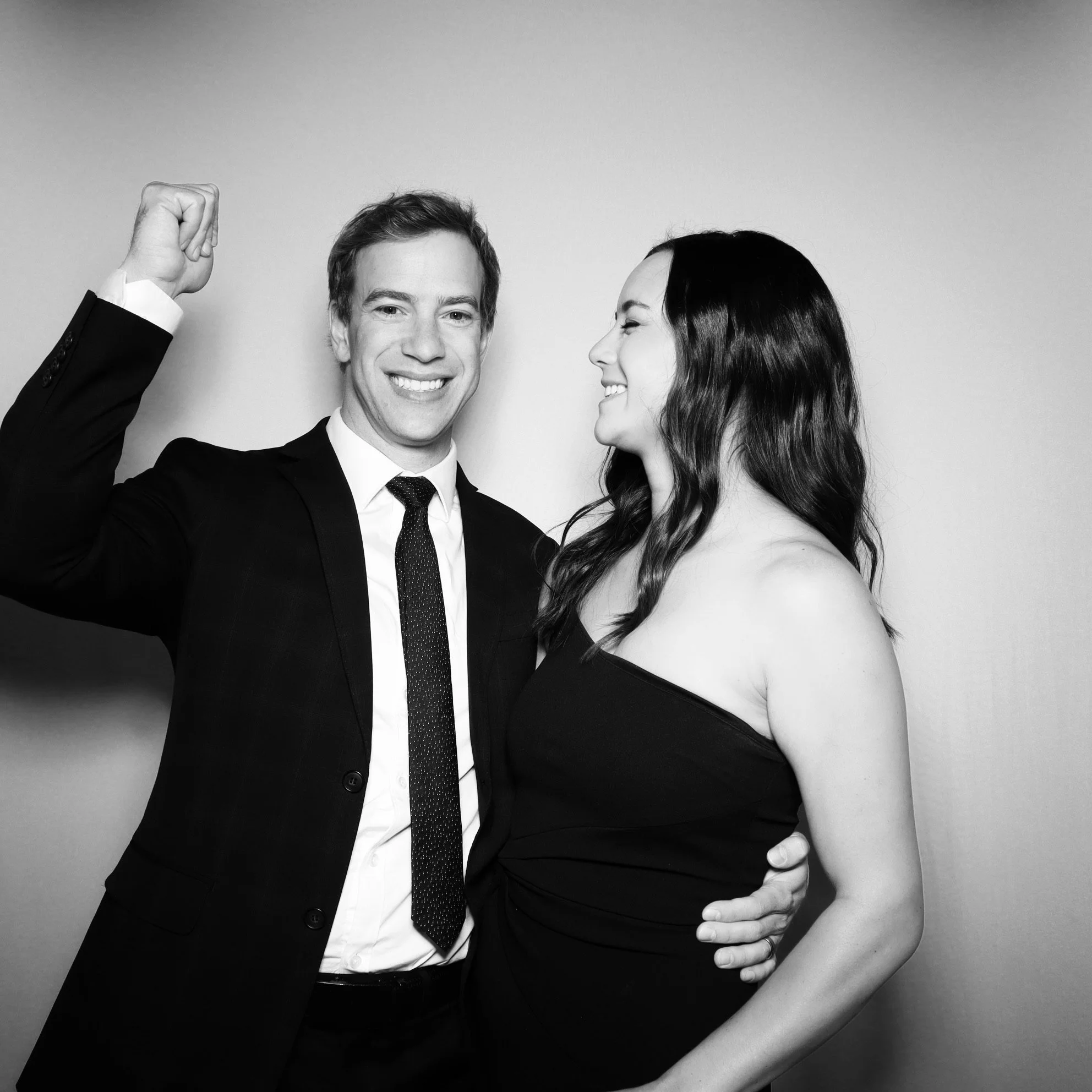 A black and white photo of a man in a suit with a tie, smiling and raising his fist, standing next to a pregnant woman in a strapless dress, both smiling.