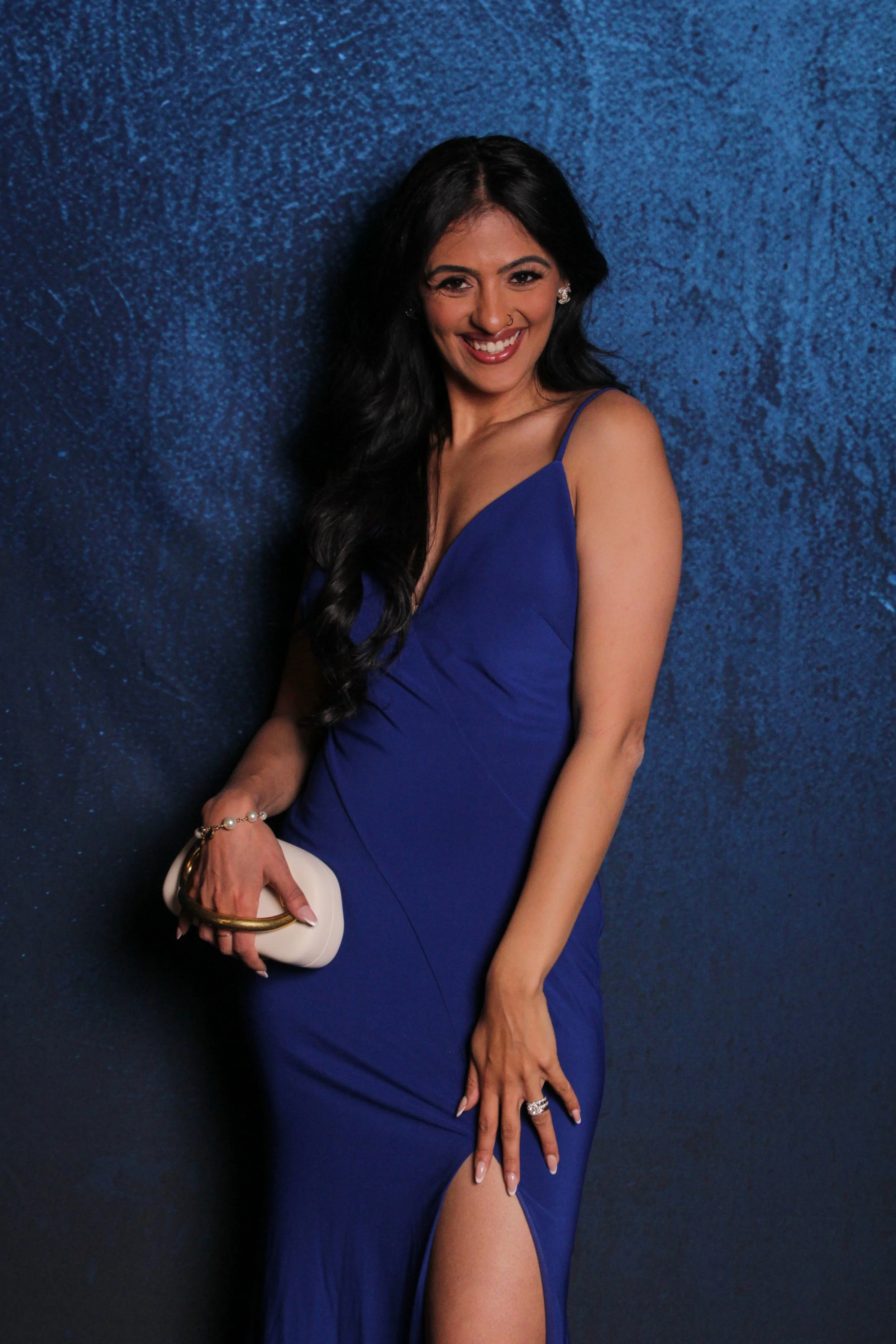 A woman with long black hair, smiling, wearing a blue dress with spaghetti straps and a slit in the skirt, holding a white clutch purse with a gold handle, standing against a textured blue background.