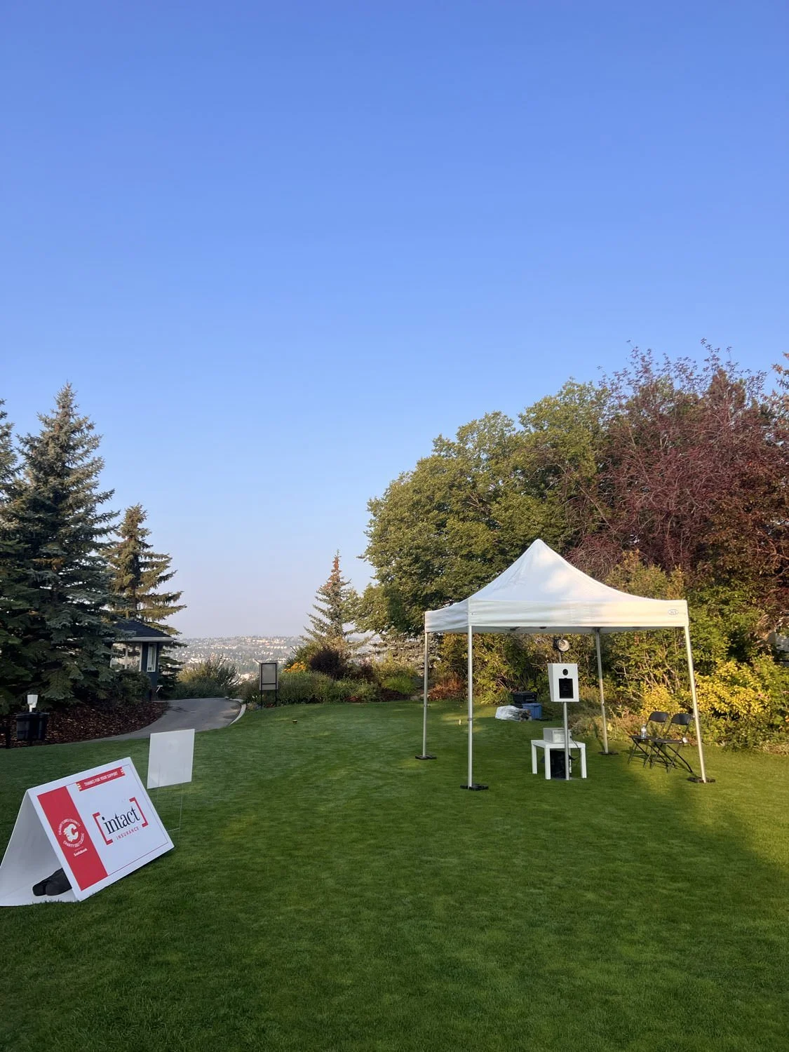 Outdoor photo booth setup with a white canopy tent, chairs, and a sign on the grass, surrounded by trees and a clear blue sky.