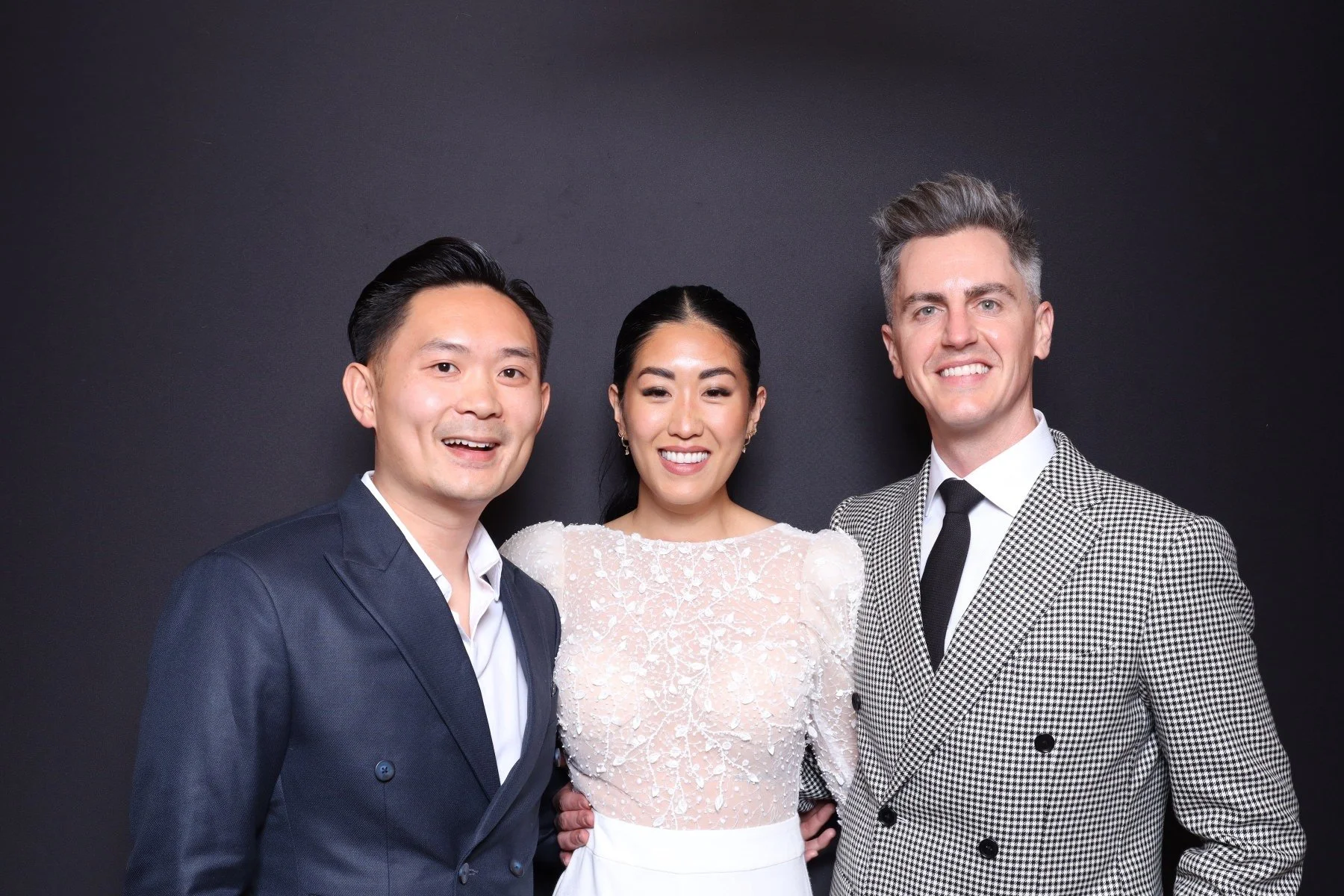 Three well-dressed people, two men and one woman, smiling and standing together against a black background. The woman is in the center wearing a white dress with lace details, and the men are on either side wearing suits, the man on the left in a dar