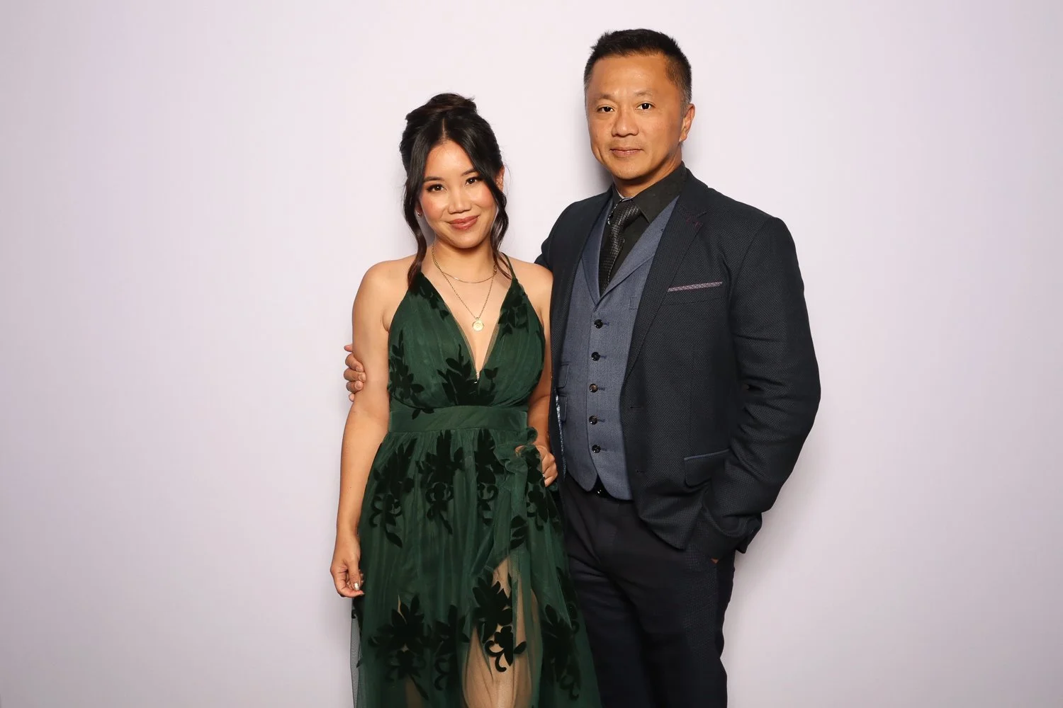 A woman in a green dress and a man in a dark suit standing together against a white background.