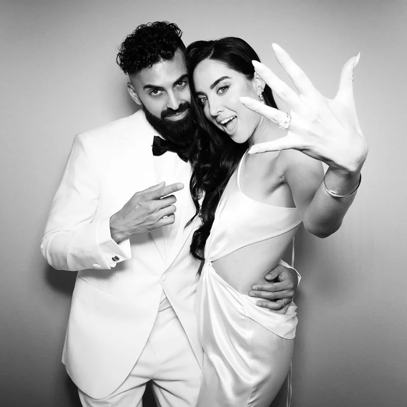 A man in a tuxedo and a woman in a dress posing together for a black and white photo, with the woman extending her hand towards the camera and smiling.