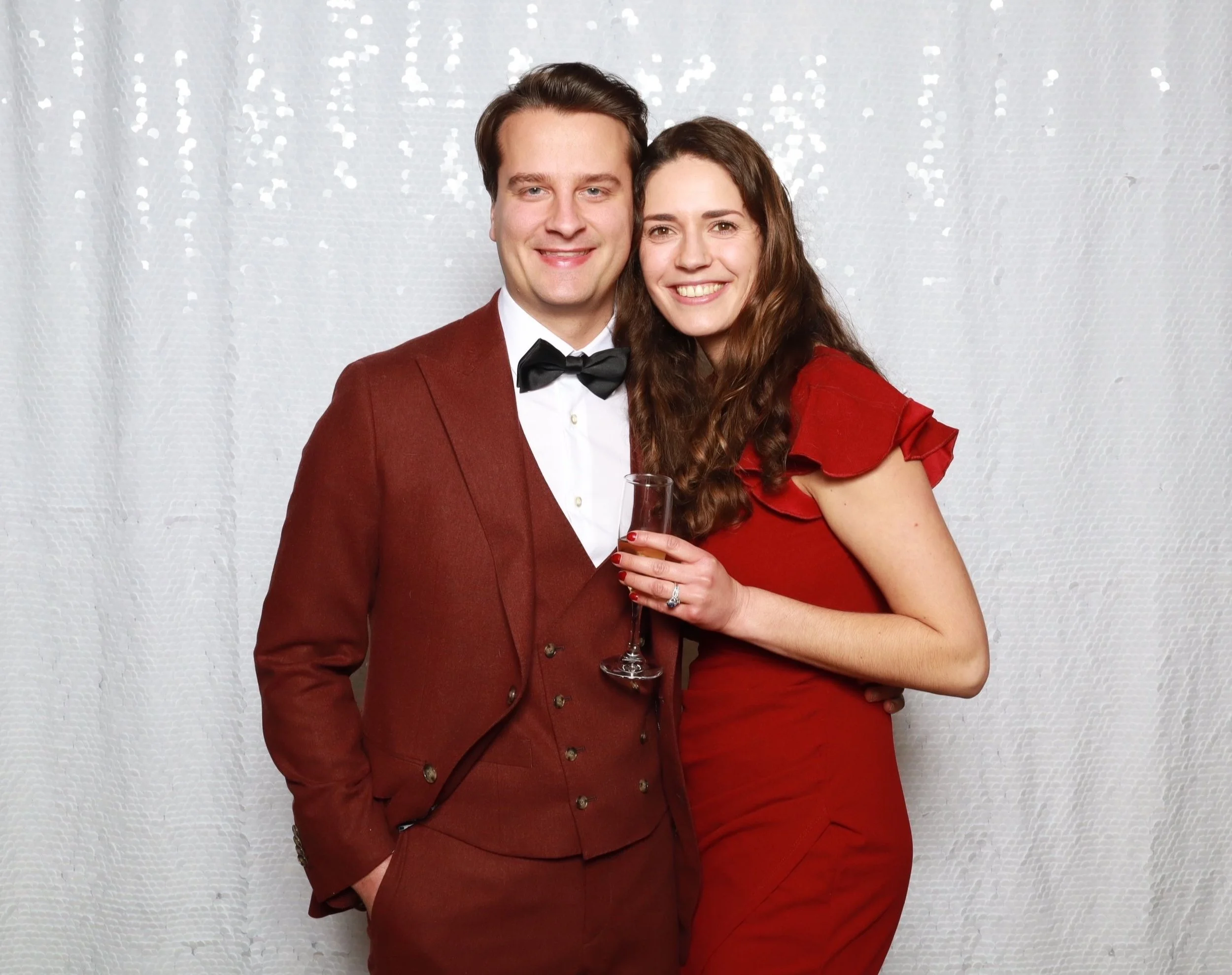 A smiling man and woman dressed in elegant attire, standing close together in front of a light-colored, textured backdrop. The man is wearing a brown suit with a black bow tie, and the woman is wearing a red dress and holding a glass of champagne.