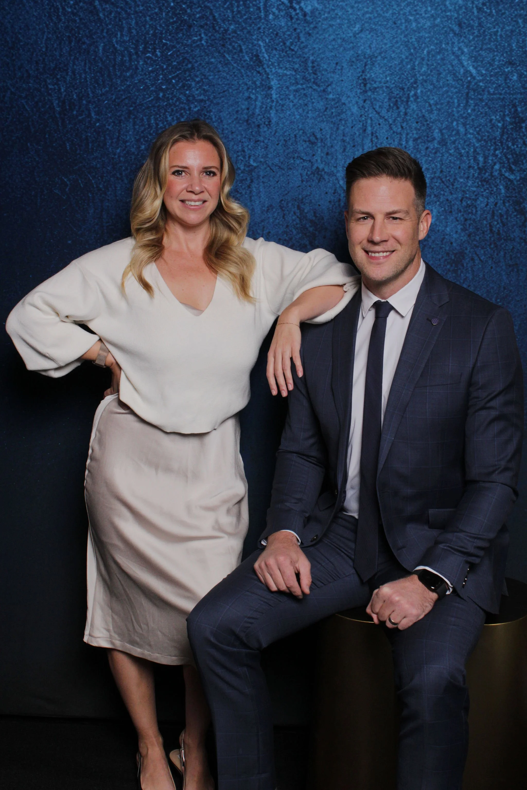 A woman with blonde, wavy hair wearing a cream-colored blouse and skirt, standing with her arm resting on a seated man in a dark suit and tie, both smiling, against a textured blue background.
