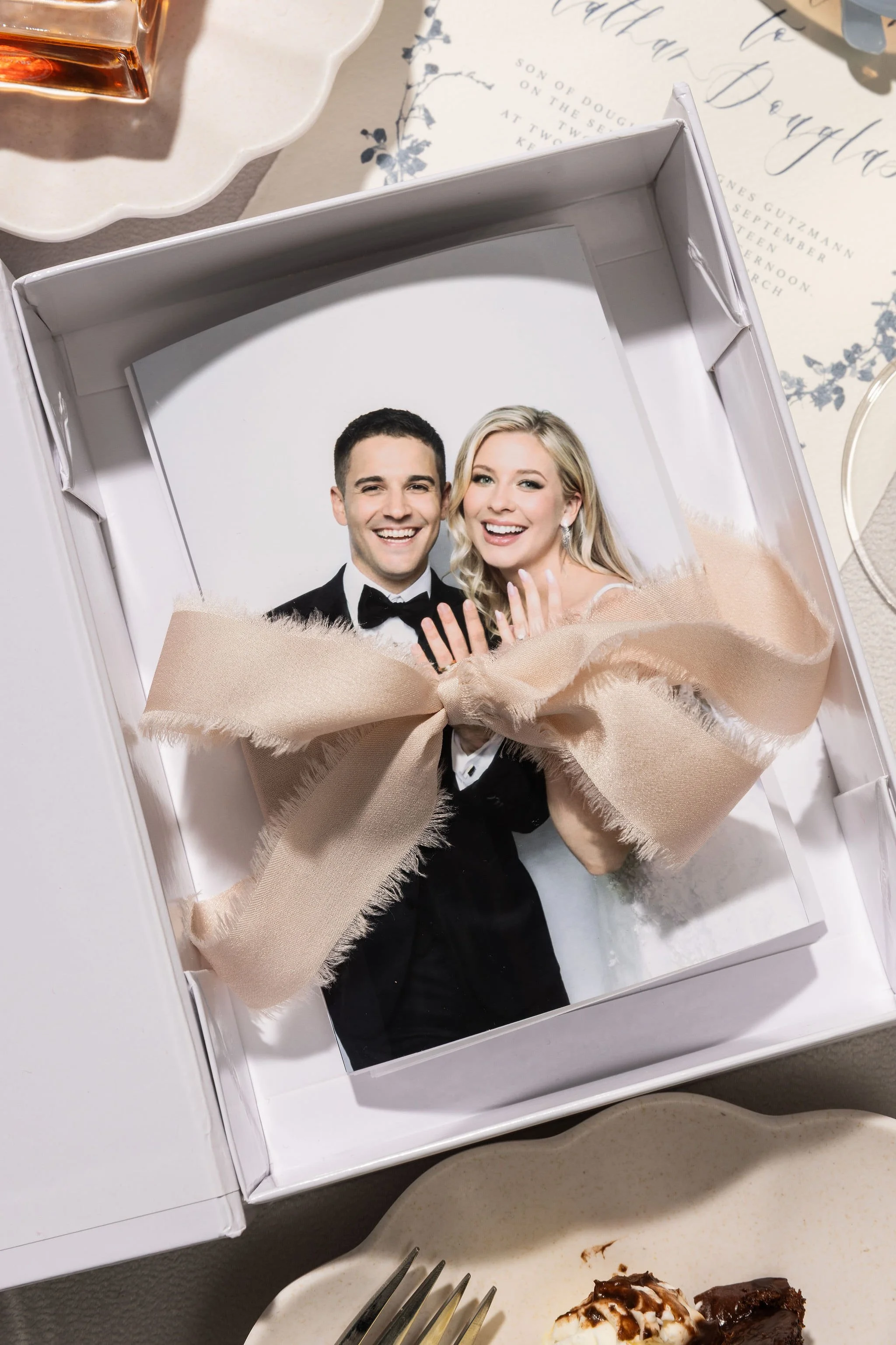 A photo of a smiling bride and groom in their wedding attire, displayed in a white box with a beige ribbon, placed on a table with wedding invitations and desserts nearby.