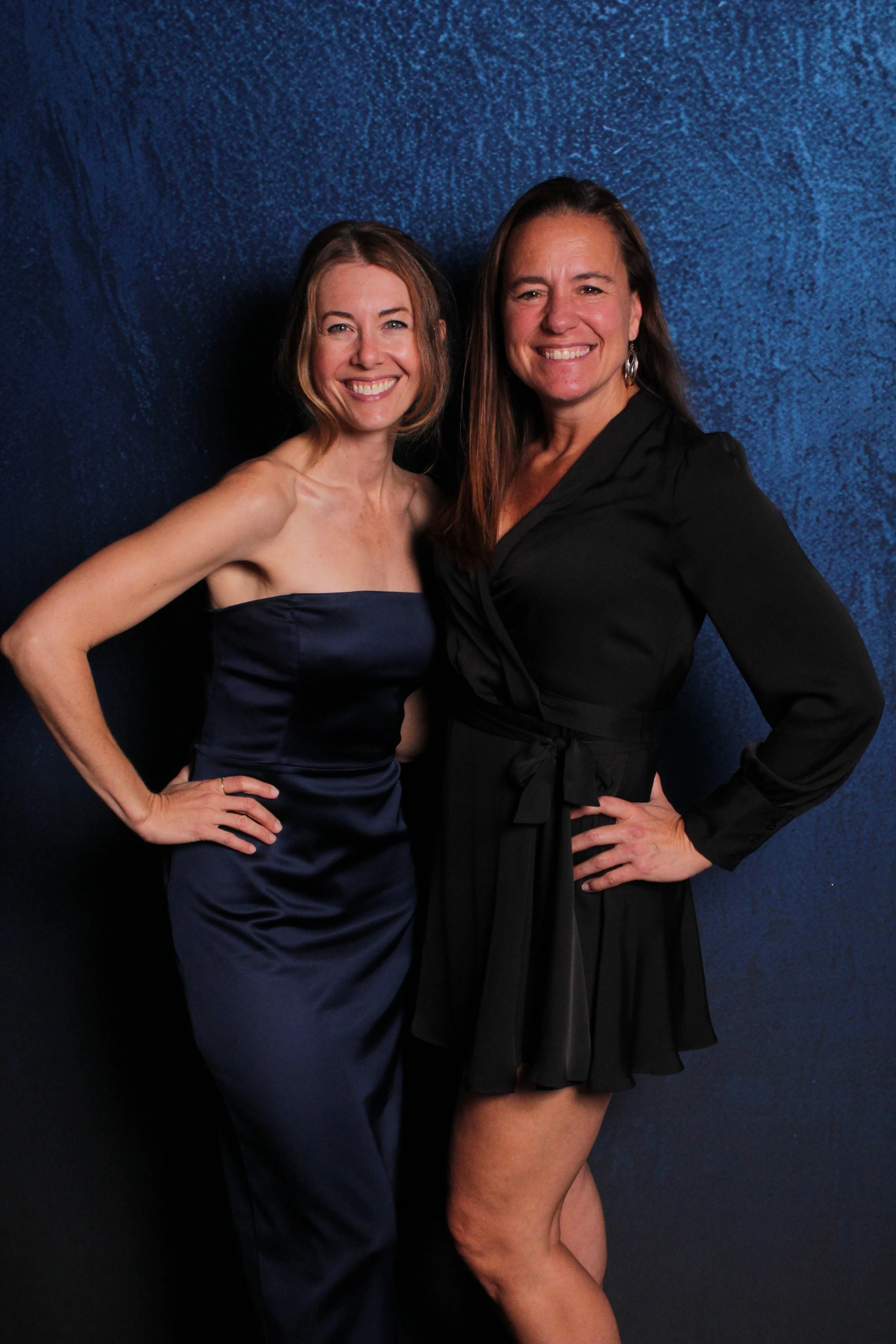 Two women smiling and posing for a photo against a dark blue background. The woman on the left wears a navy blue strapless dress, and the woman on the right wears a black dress with long sleeves and a waist tie.