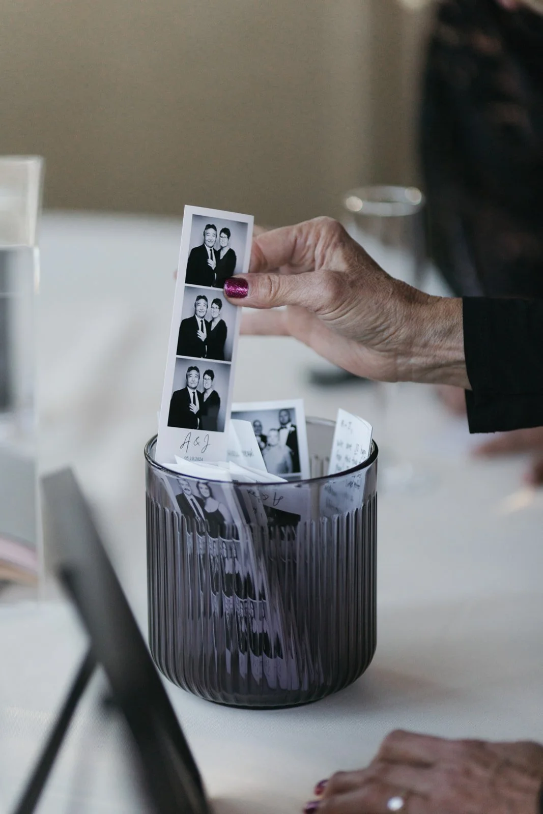 A hand with red glitter nail polish is holding a black-and-white photo strip of three pictures, placed in a clear ribbed glass vase on a white table, with more photo strips and handwritten notes inside the vase.