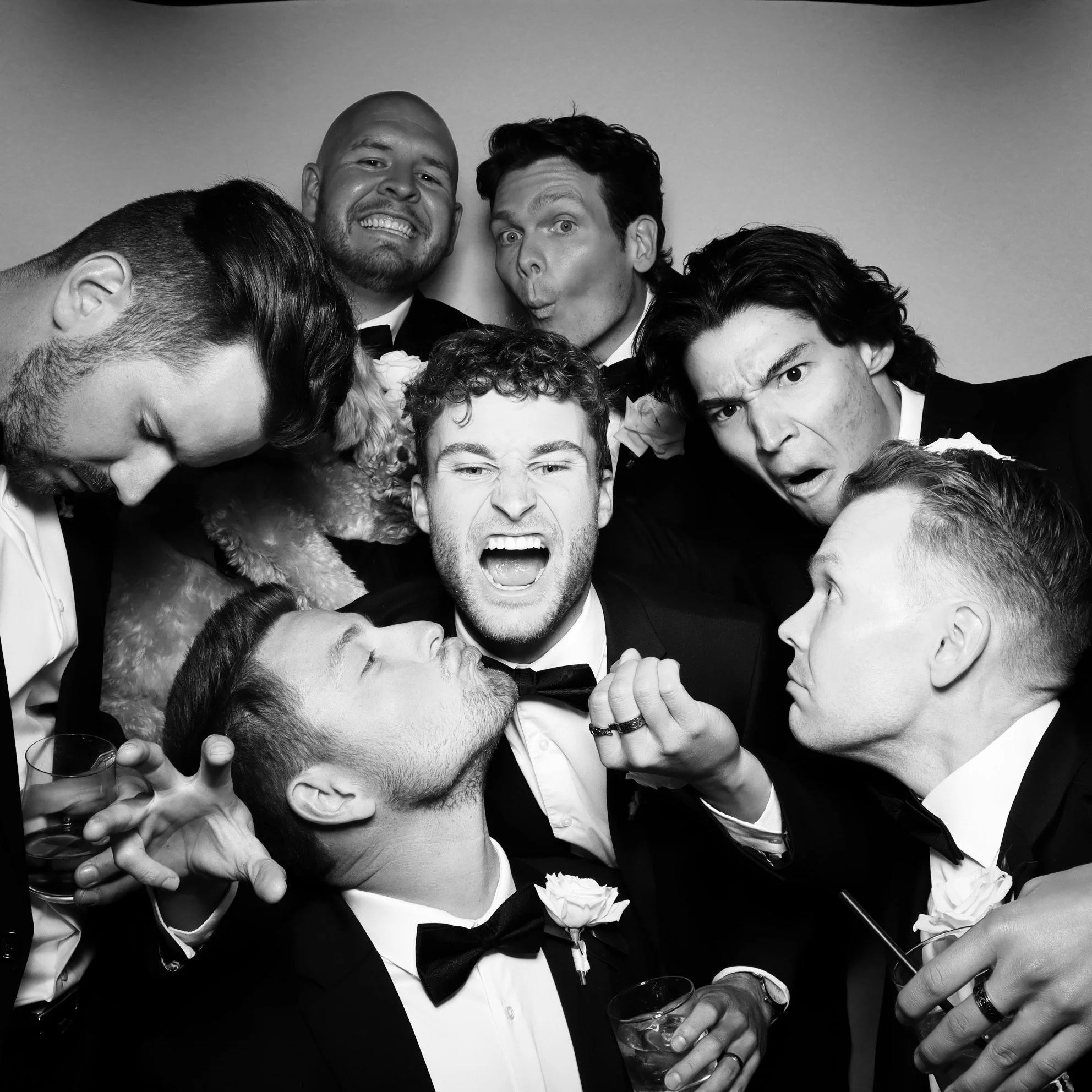 Black and white photo of a group of men in tuxedos making humorous and expressive faces, some holding drinks, with one man at the center yelling or singing, and a small dog among them.