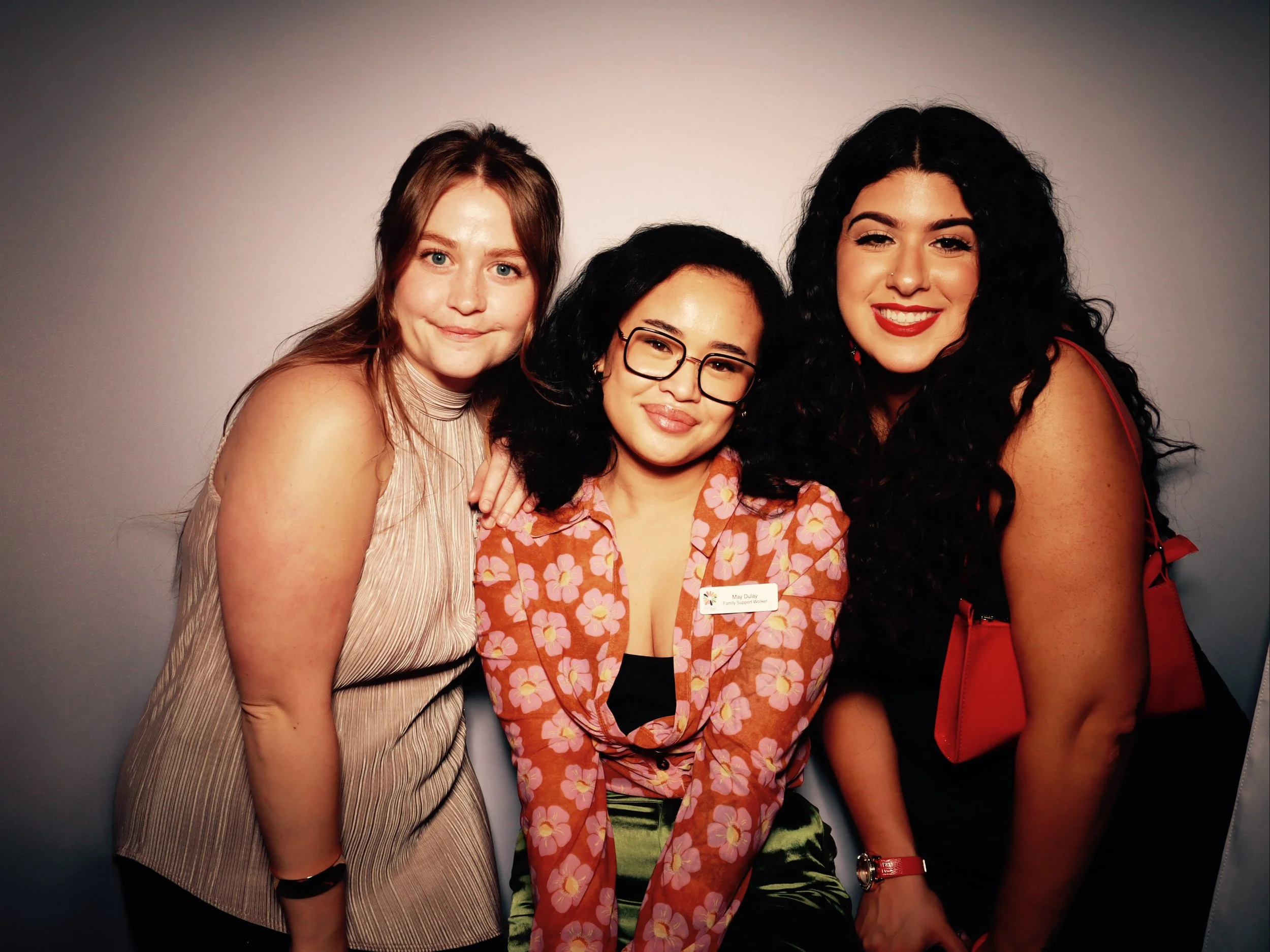 Three women smiling and posing together against a plain backdrop, one wearing glasses and a floral shirt, another with long curly hair and red lipstick, and the third with light brown hair and a sleeveless beige top.