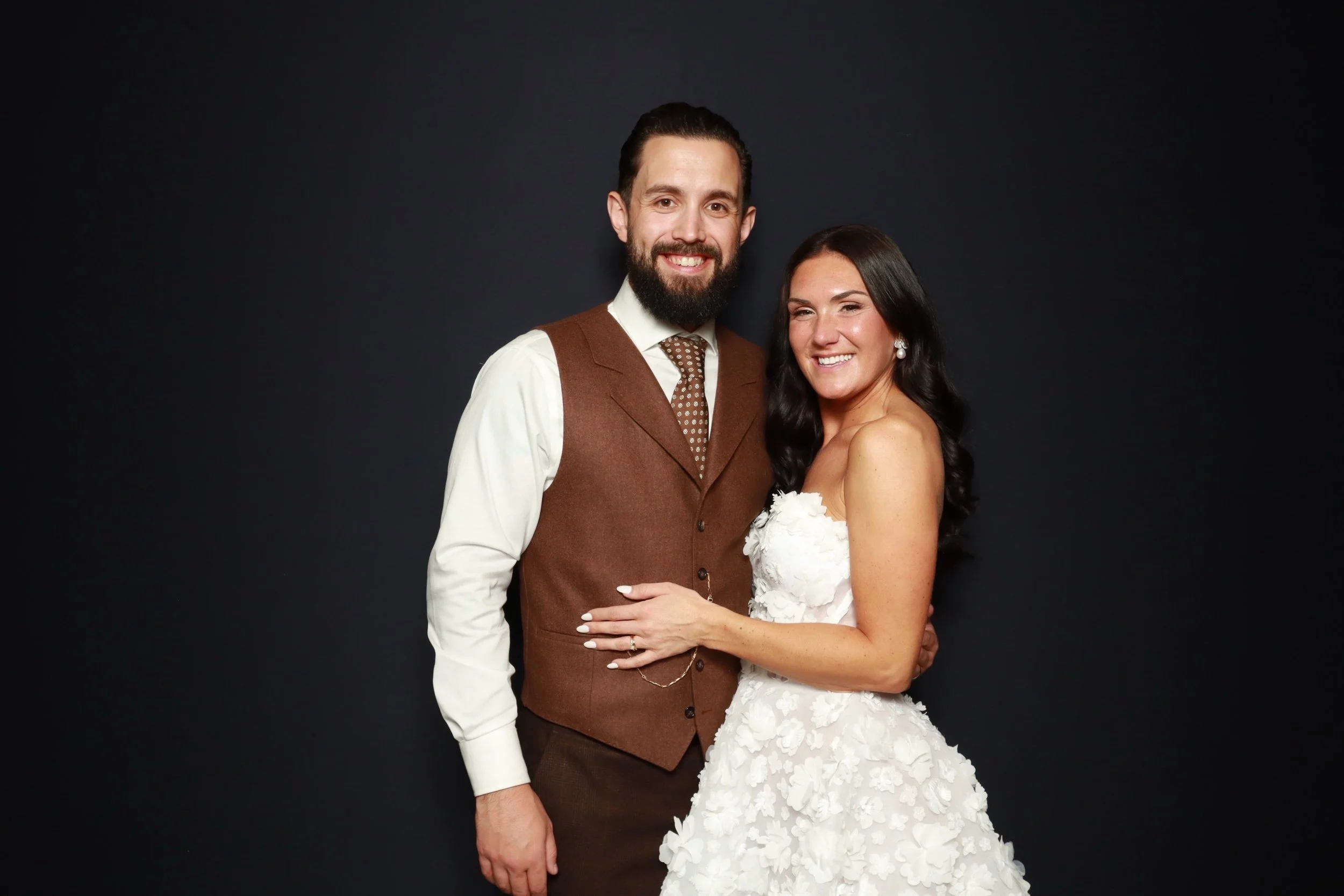 a color photo from a Flash Co. wedding photo booth featuring a smiling bride in a white floral dress and a groom in a brown vest and tie, posing together against a solid black backdrop.