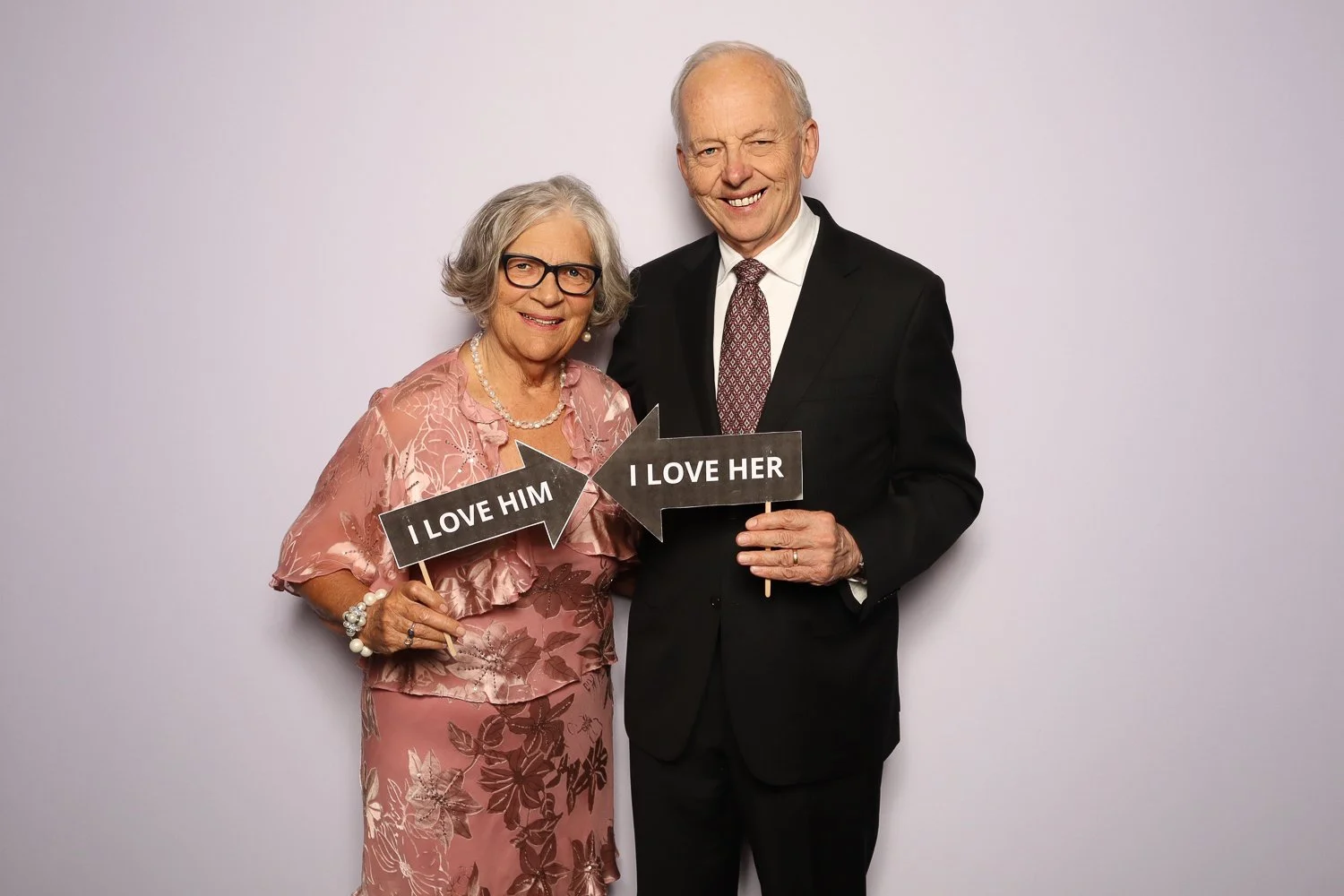 An elderly woman in a pink floral dress and glasses, and an elderly man in a black suit and tie, standing together holding signs that say "I LOVE HIM" and "I LOVE HER," smiling against a plain background.