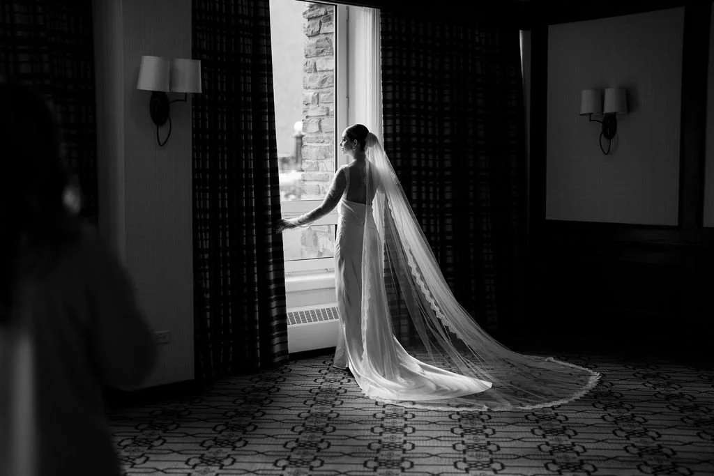 A bride in a wedding dress and veil stands by a window looking outside, with her hand on the curtain, in a dimly lit room.