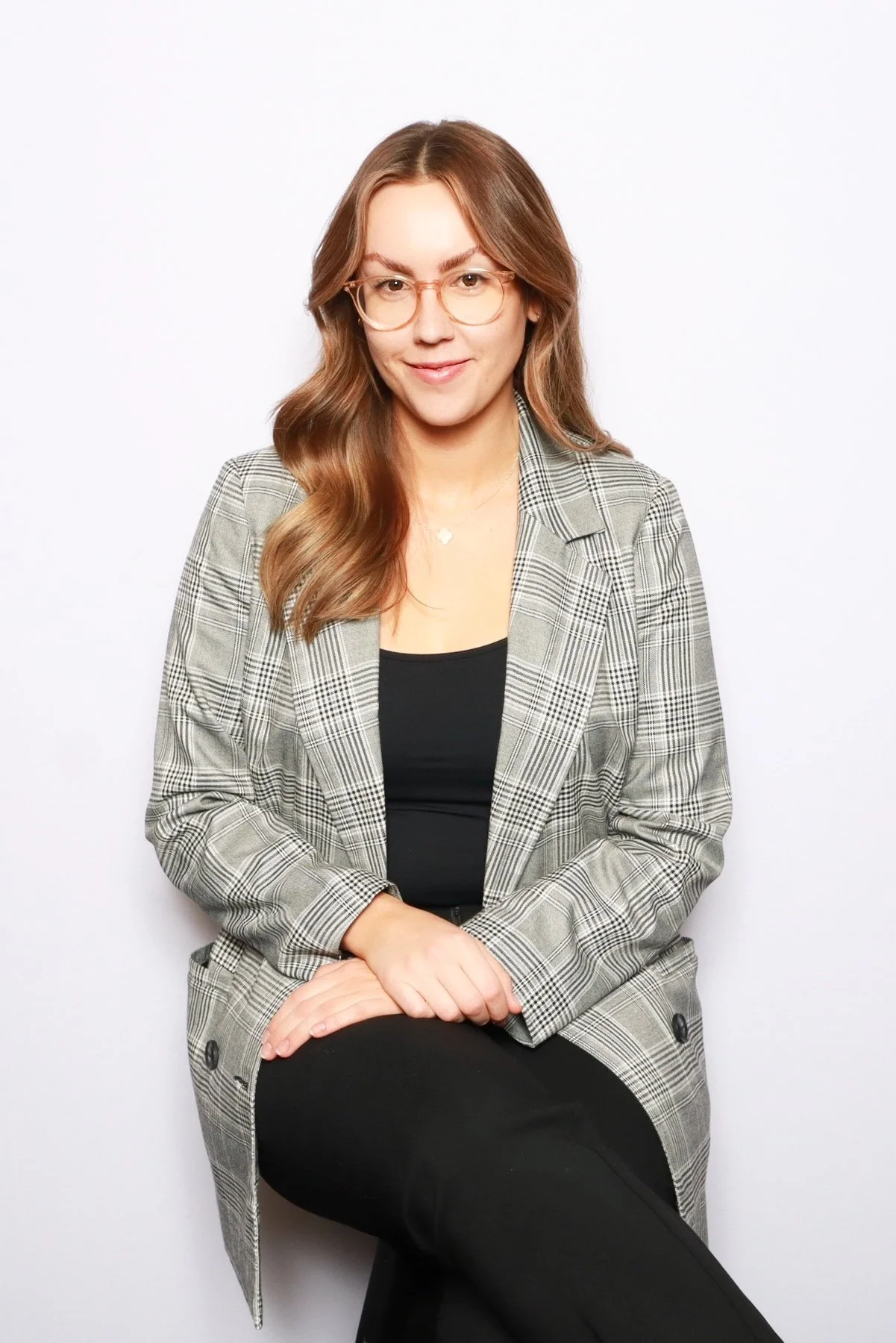A woman with long wavy brown hair, wearing glasses, a black top, and a plaid blazer, sitting against a white background.