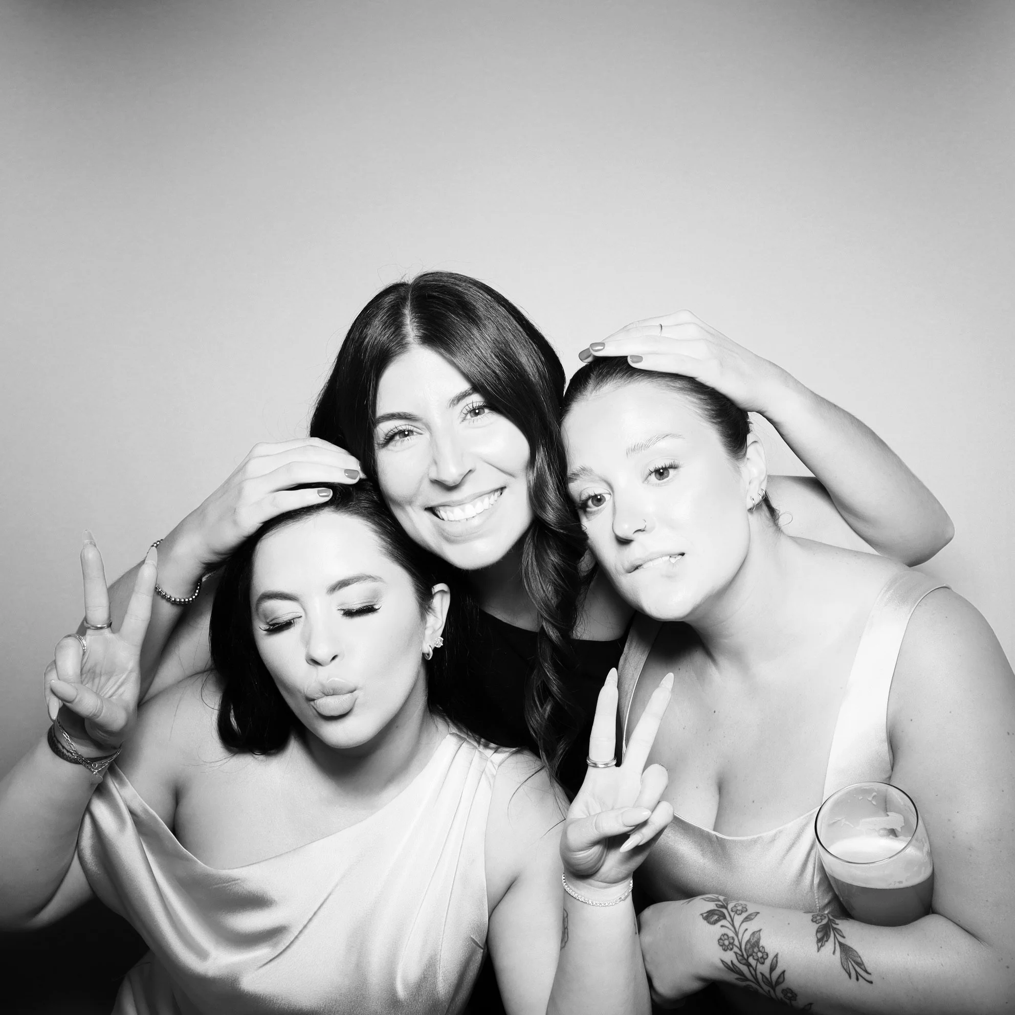 Black and white photo of three women smiling and making peace signs, with one woman in the center, one to her left, and one to her right, against a plain background.