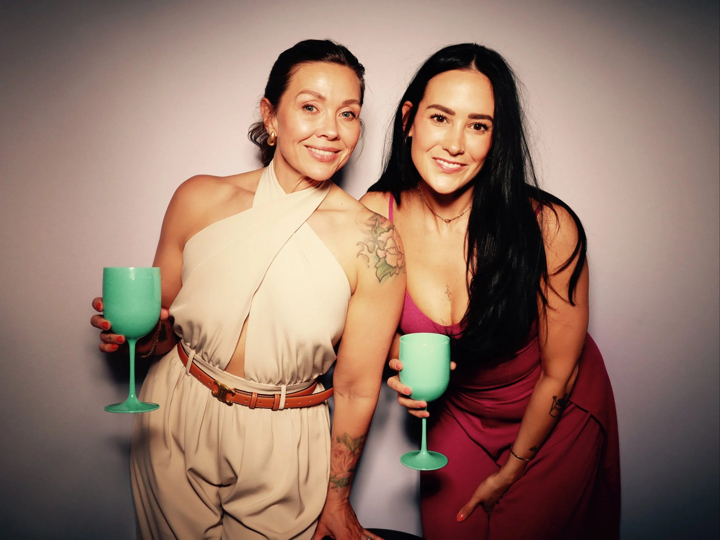 Two women smiling and holding green cups, standing close together against a plain backdrop.