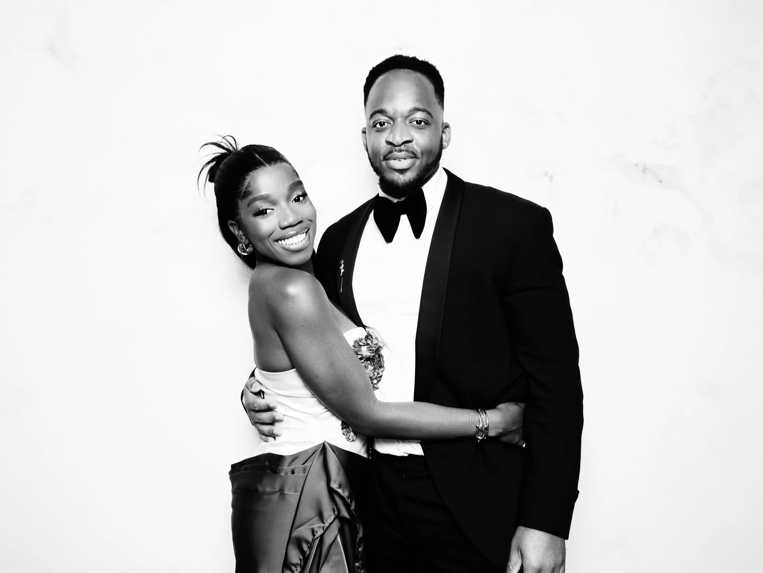 A smiling woman in a strapless dress and a man in a tuxedo pose together against a plain background.