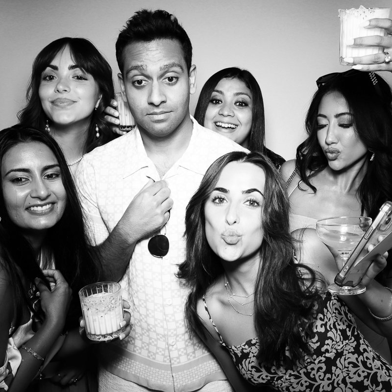 Group of six young adults at a party, posing for a photo with drinks, making playful and smiling expressions, in black and white.