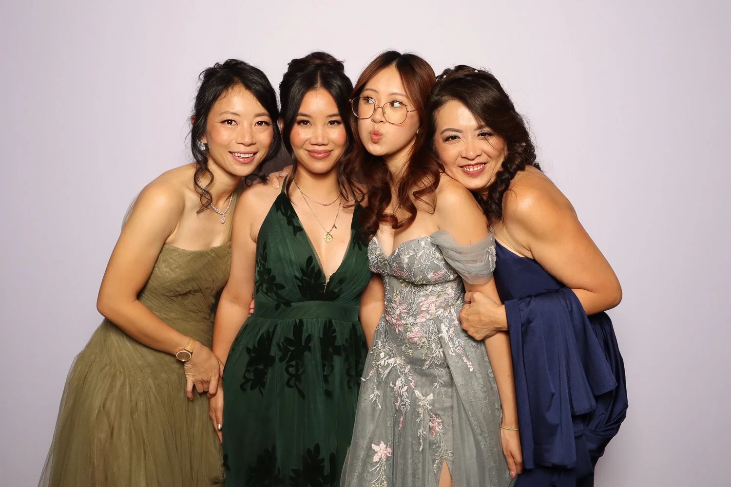 Group of four women dressed in formal dresses, standing together and smiling, against a plain light background.