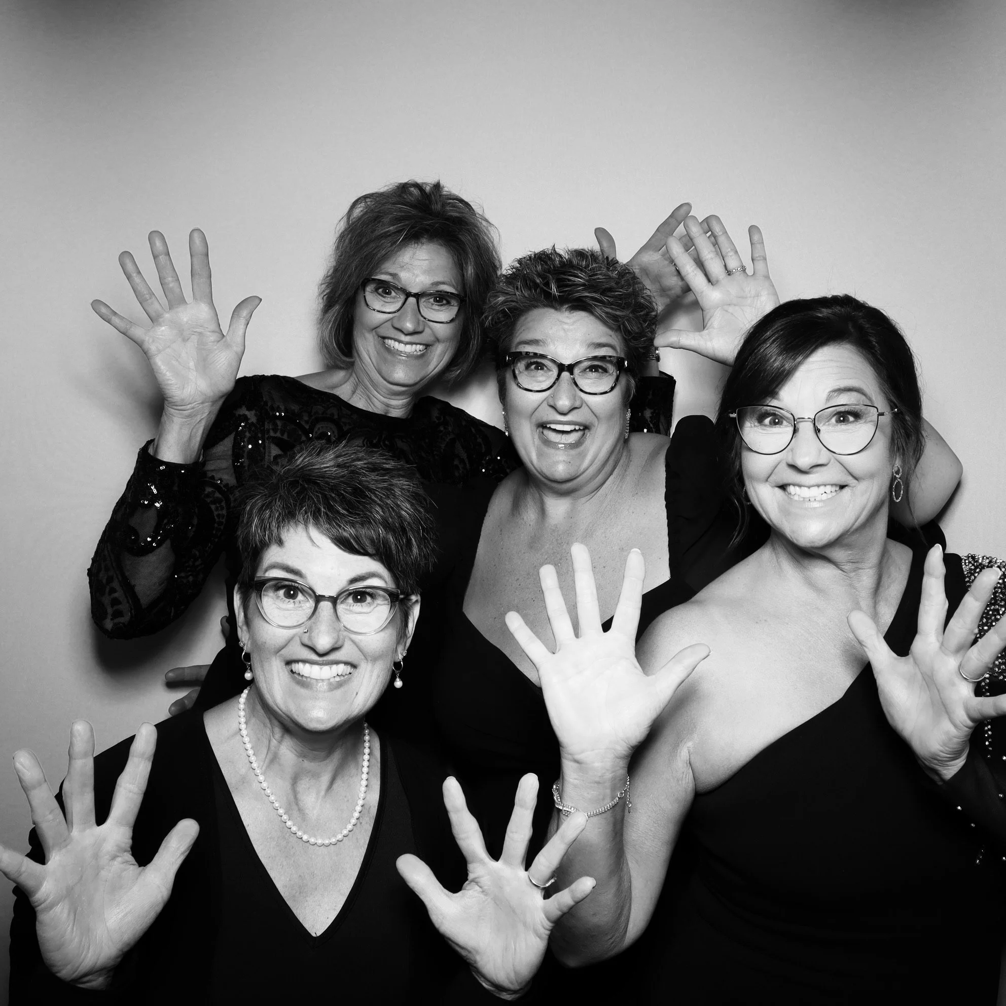 Five women smiling and waving at the camera, wearing black dresses and glasses, posed against a plain background.