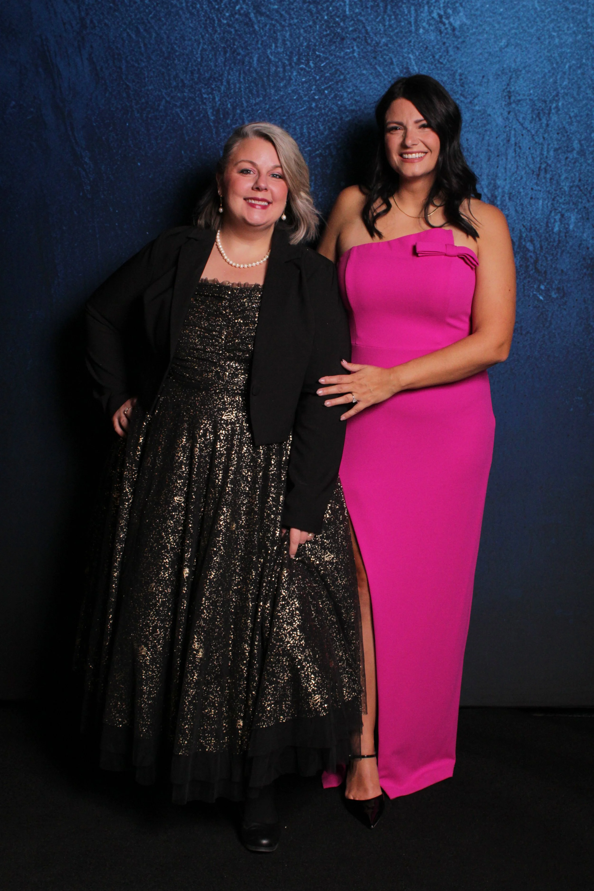 Two women standing against a dark blue textured wall, dressed in formal evening attire, smiling at the camera.