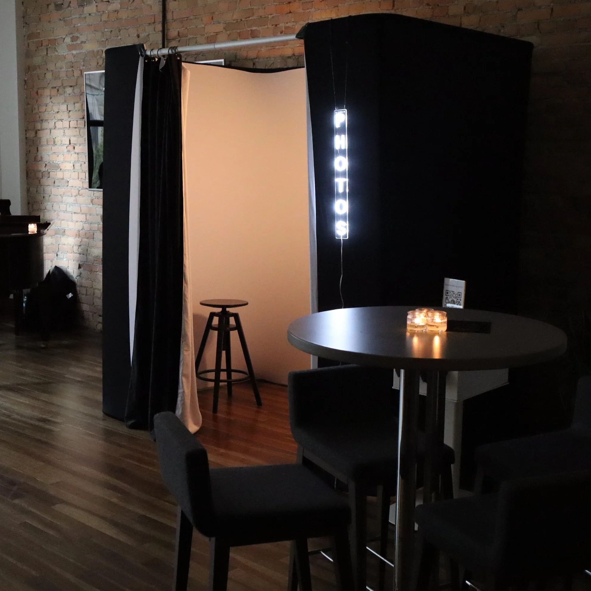 Photo booth with black curtains, a beige backdrop, a small stool, a round table with candles, and a lit sign spelling 'PHOTOS' in a dimly lit room with wooden floors and a brick wall.