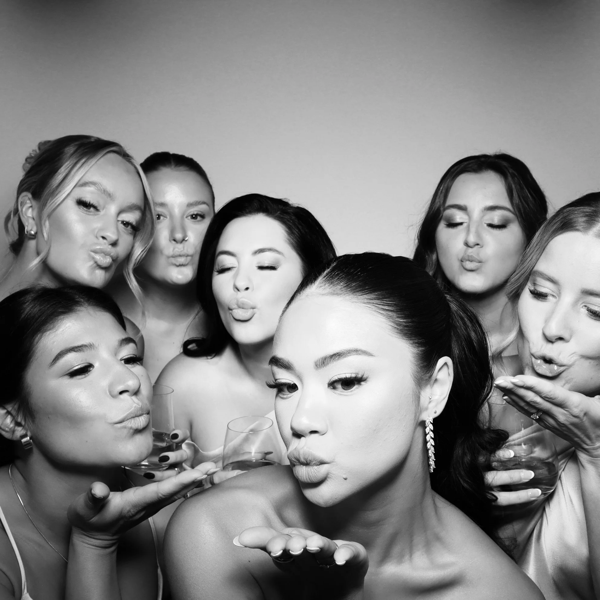 Black and white photo of eight women blowing kisses, holding glasses of champagne, dressed elegantly, with one woman in the foreground and the others behind her.