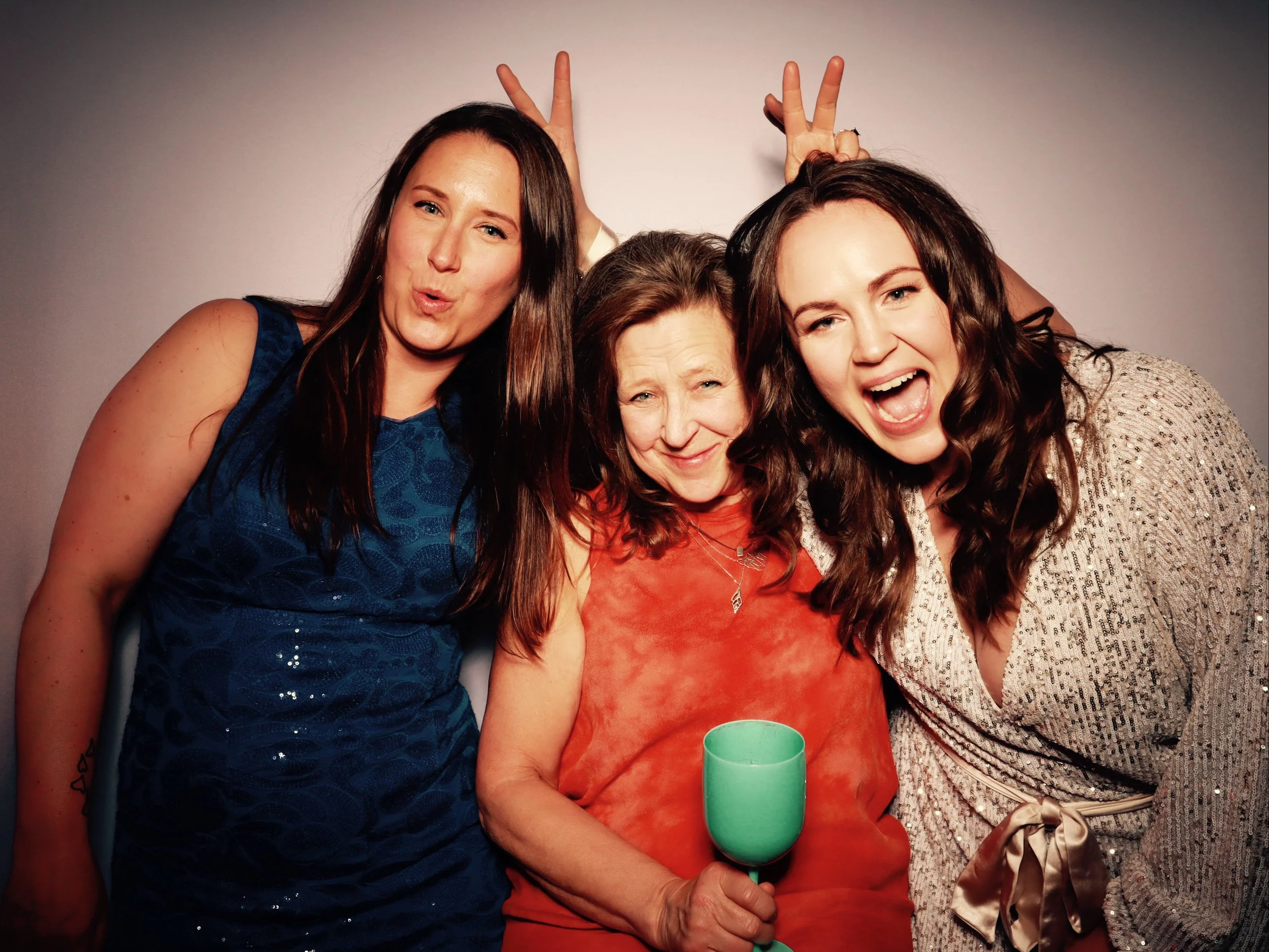 Three women celebrating, one holding a drink, cheerful expressions, playful poses with hand gestures, festive attire, neutral background.