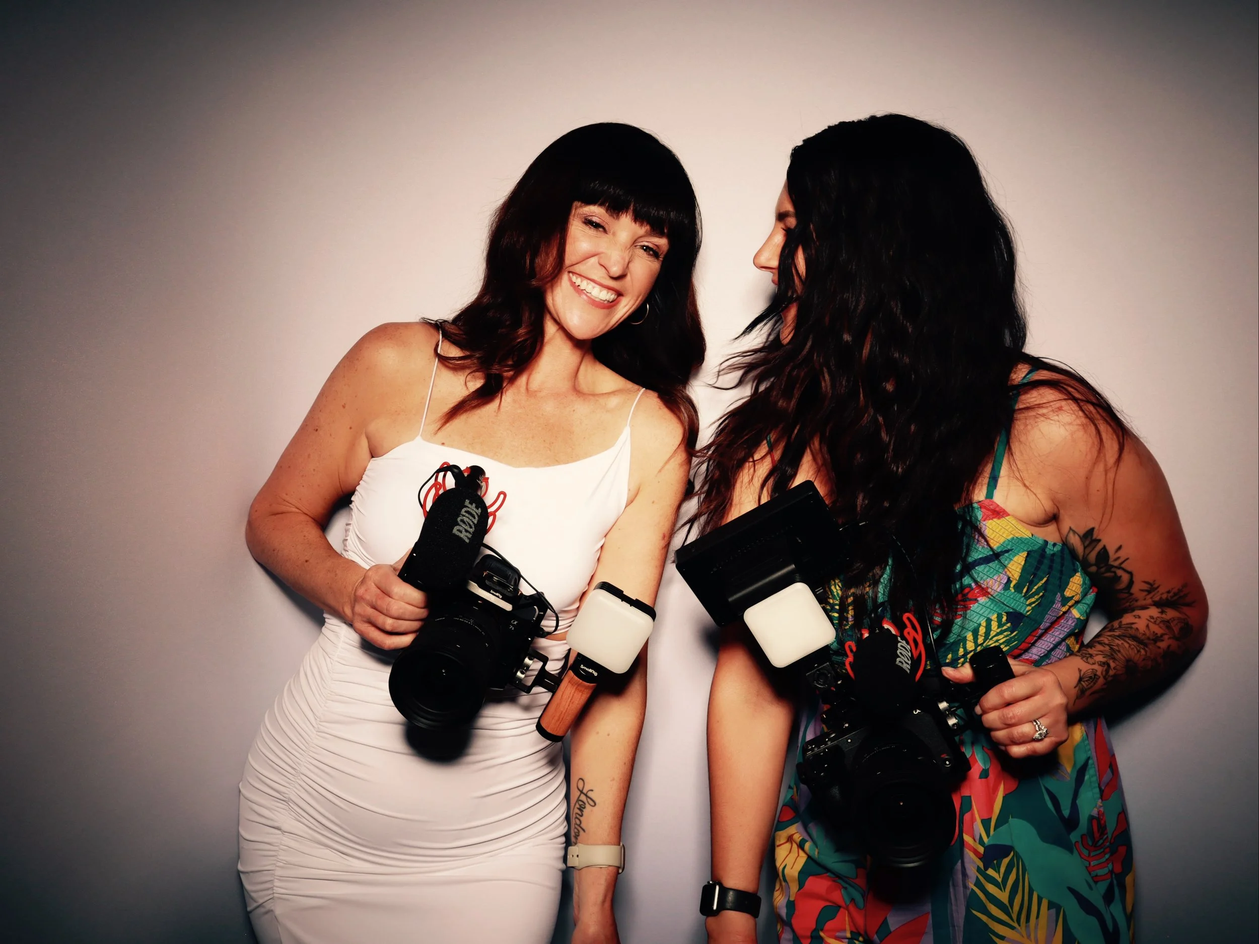 Two women smiling and holding cameras stand against a plain wall, sharing a joyful moment.