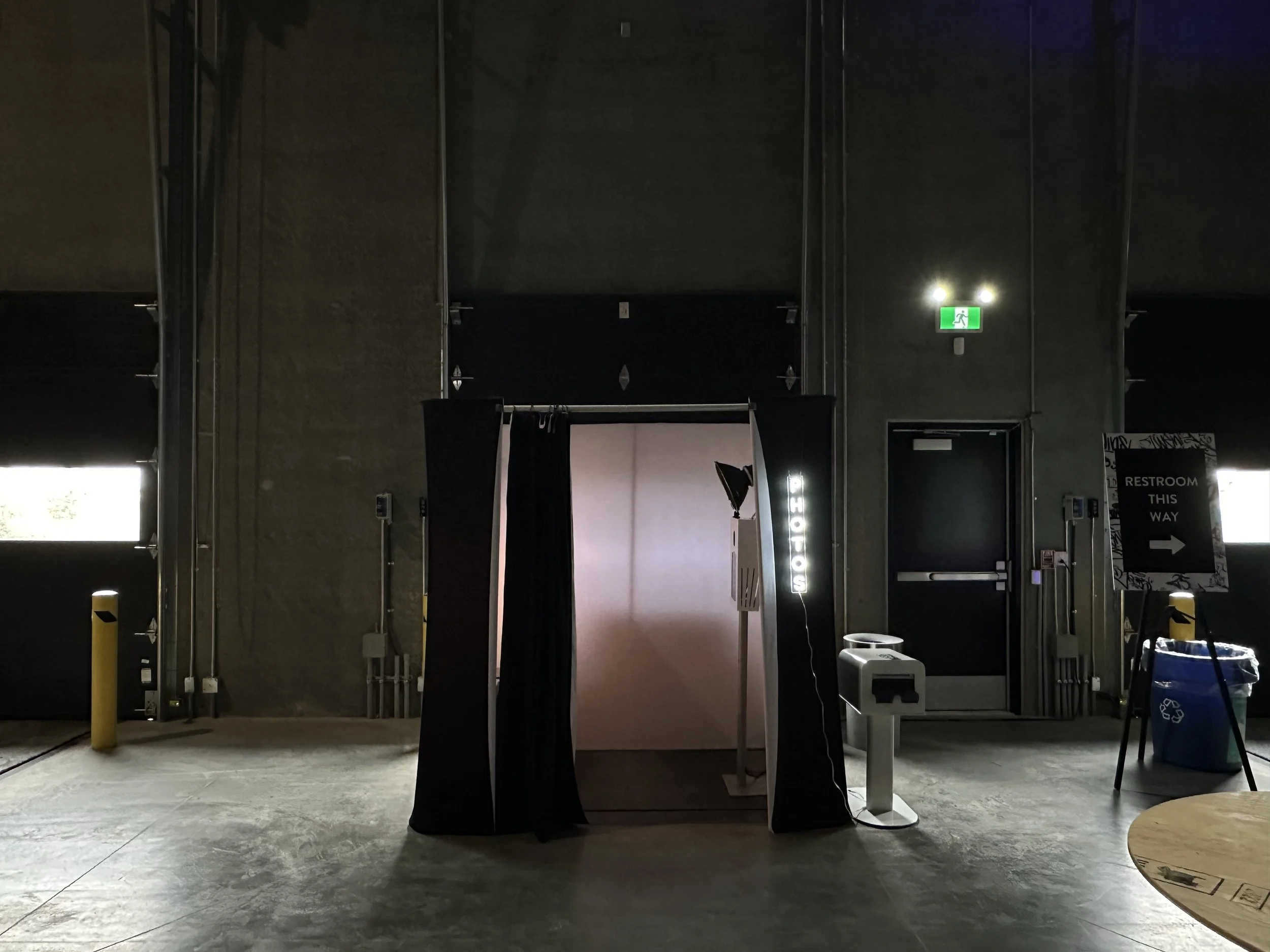 Photo booth with black curtains and a white backdrop, located in a dimly lit industrial-style space with concrete floors and dark walls. A sign on the right indicates restroom directions, and a trash can is visible nearby.