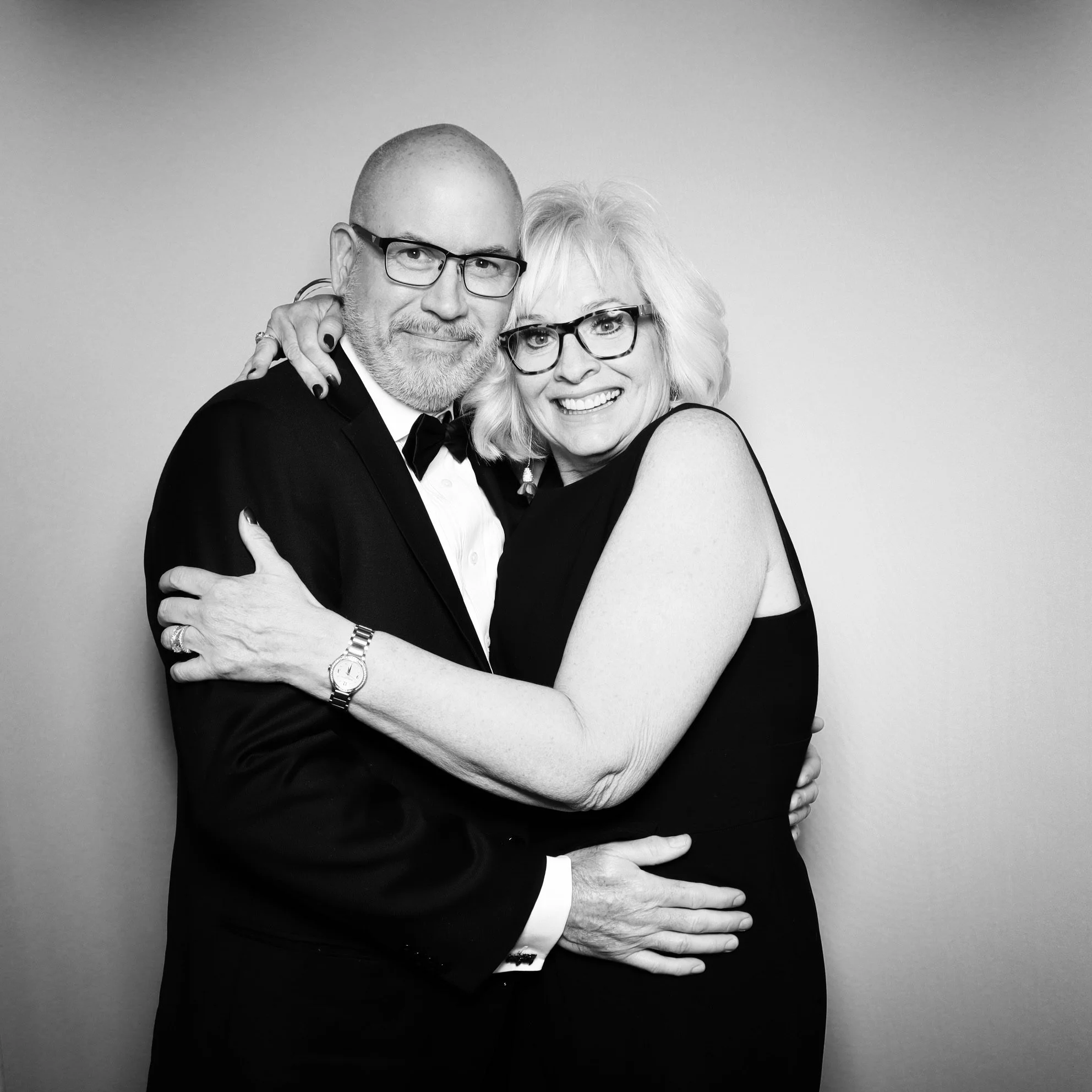 Black and white photo of a smiling man and woman embracing, both wearing glasses and formal attire, against a plain background.