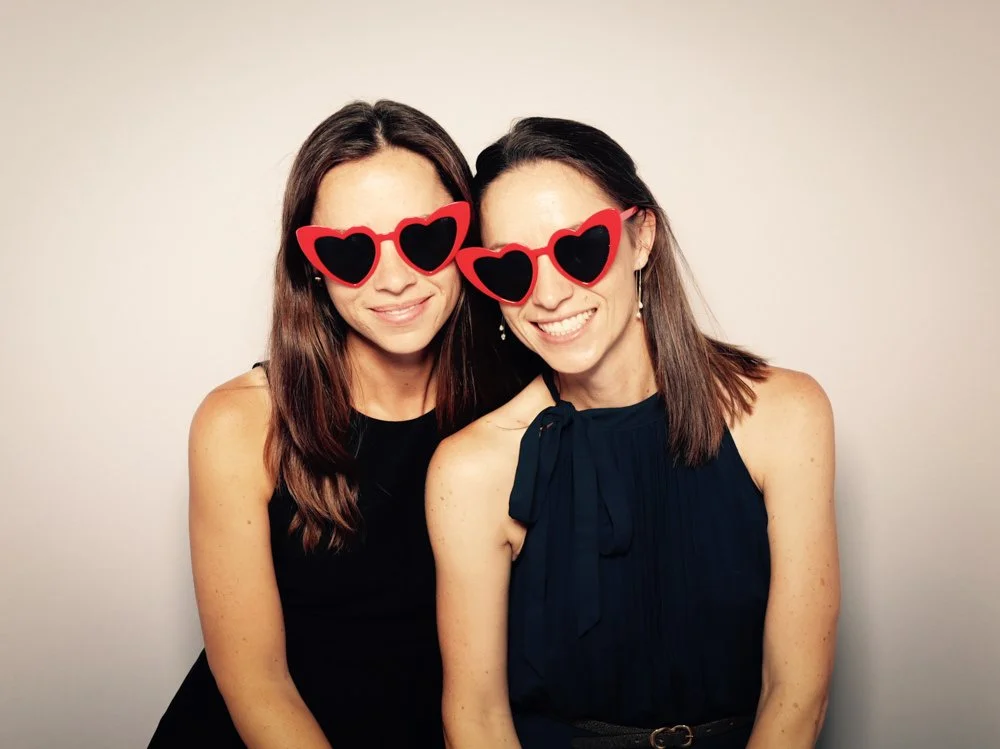 Two women smiling and wearing heart-shaped sunglasses with red frames and black lenses, standing close together against a plain white background.