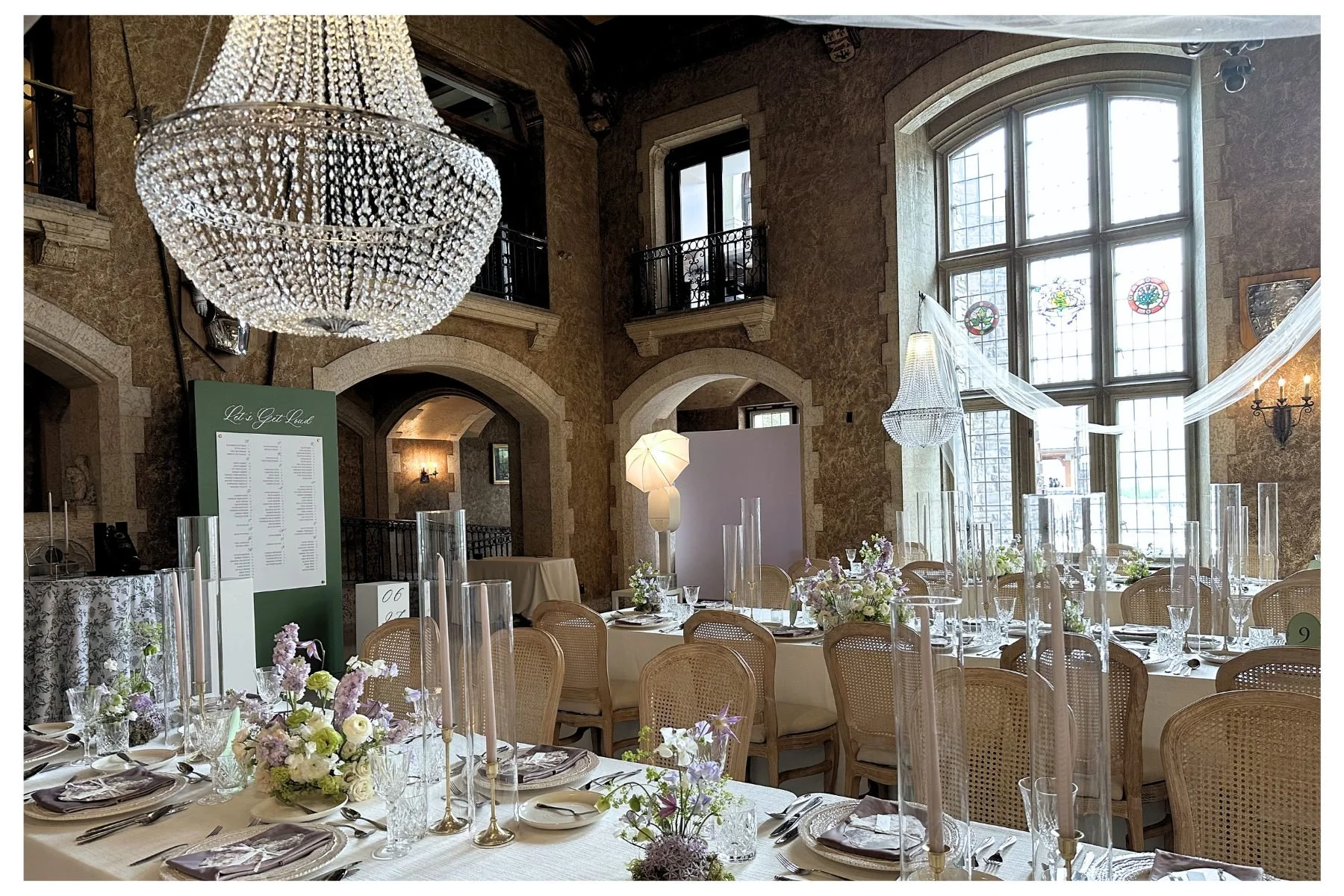 Elegant banquet hall decorated for a wedding or special event, featuring a large crystal chandelier, tall windows with stained glass, floral centerpieces, and set tables with glassware and tableware.