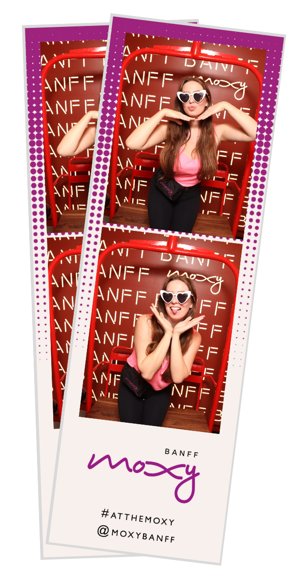 A woman in pink top and black pants with heart-shaped sunglasses posing playfully in a photo booth with a red background and the words 'Banff Moxy' repeated on the wall.