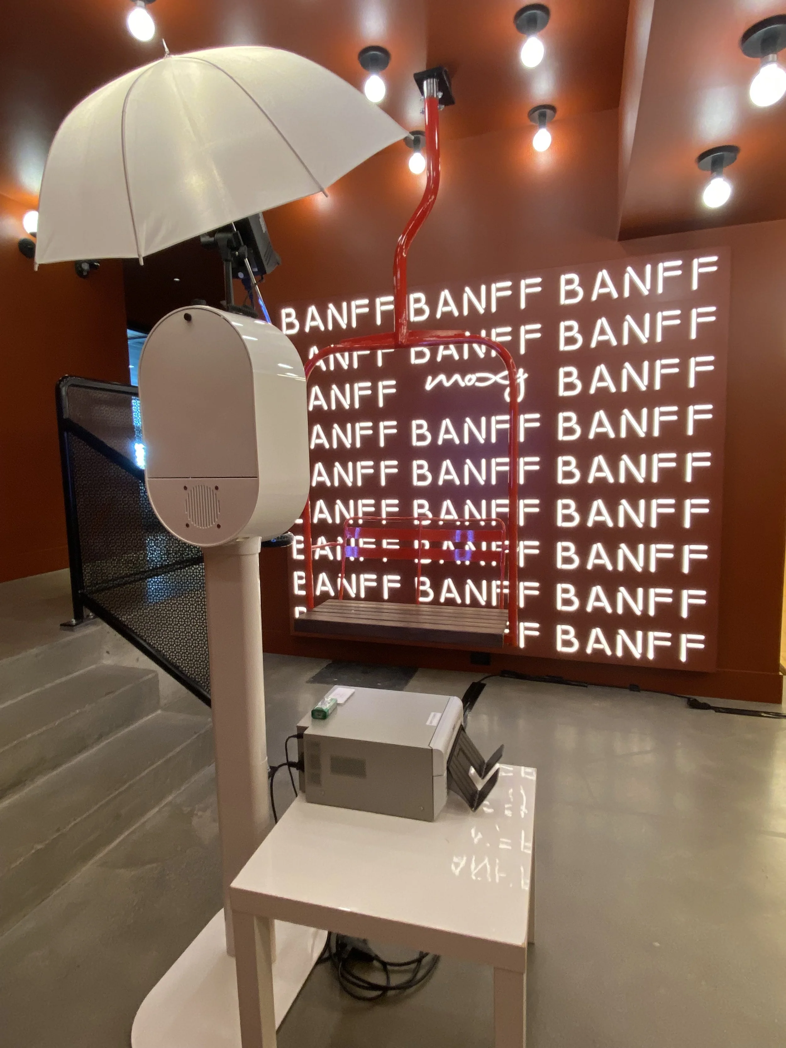 Decorative art installation with a large white umbrella, red pipe, and swing in front of a wall with repeated illuminated 'BANFF' text, on a polished floor with a staircase to the left.