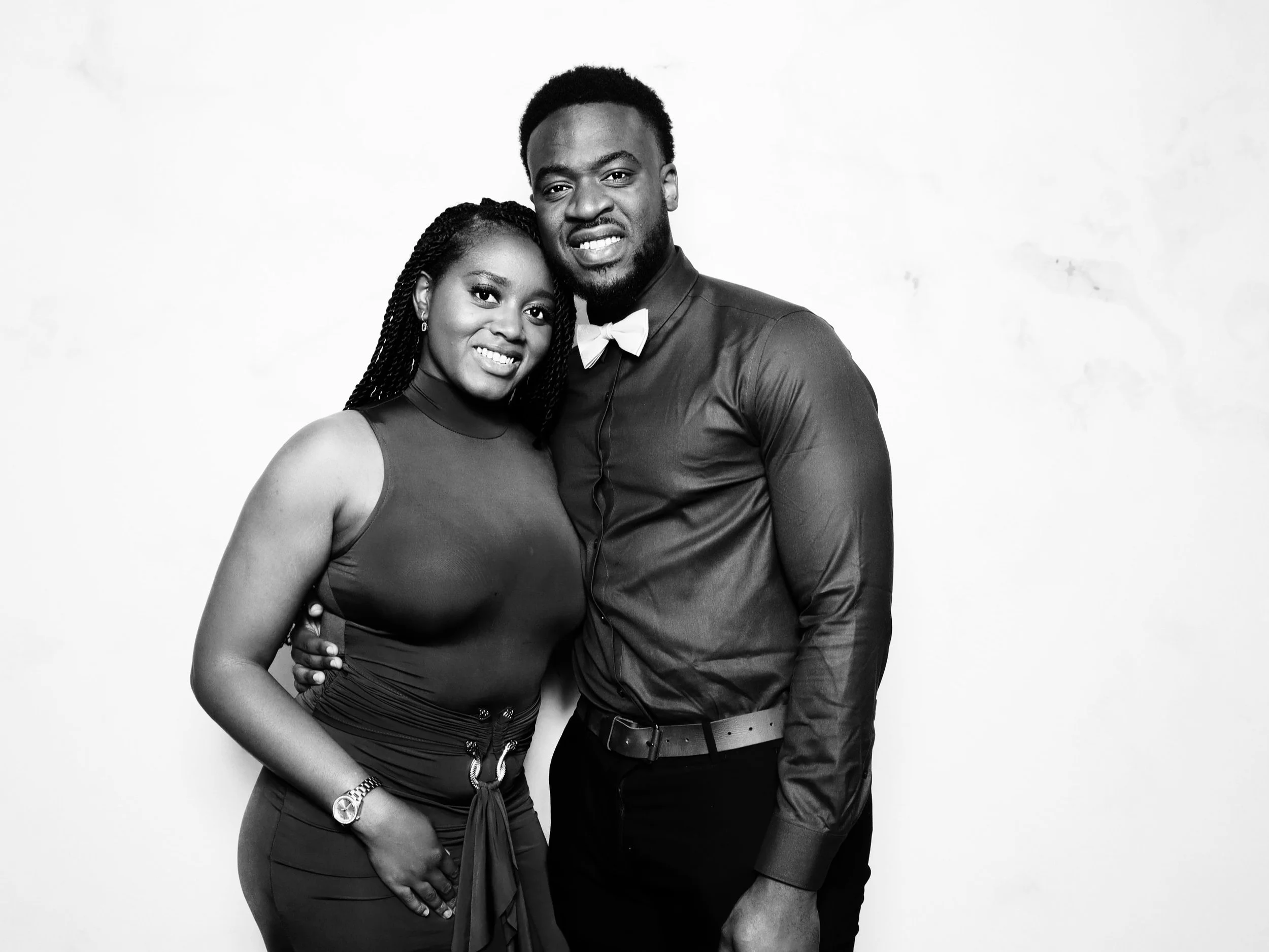 A black-and-white photo of a smiling woman and a man standing close together against a plain background, with the woman resting her hand on her hip and the man wearing a bow tie.