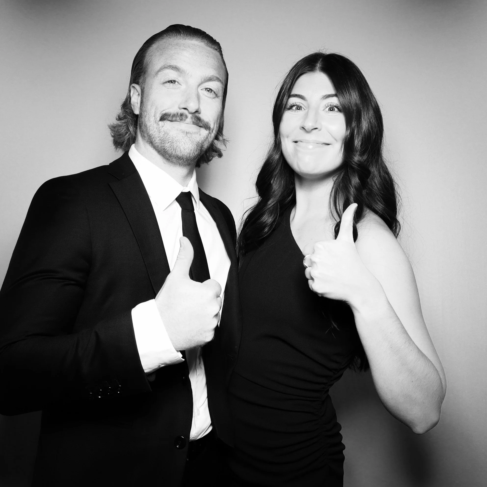 Black and white photo of a man in a suit and a woman in a dress, both giving thumbs-up signs.