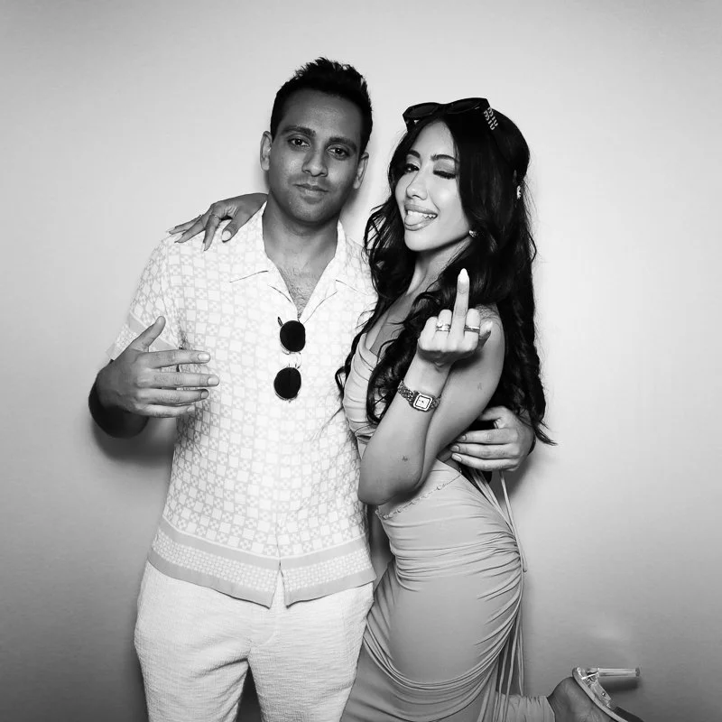 A man and woman posing together, the woman sticking her tongue out and showing middle fingers, the man with a neutral expression, both dressed casually, standing against a plain background in a black and white photo.