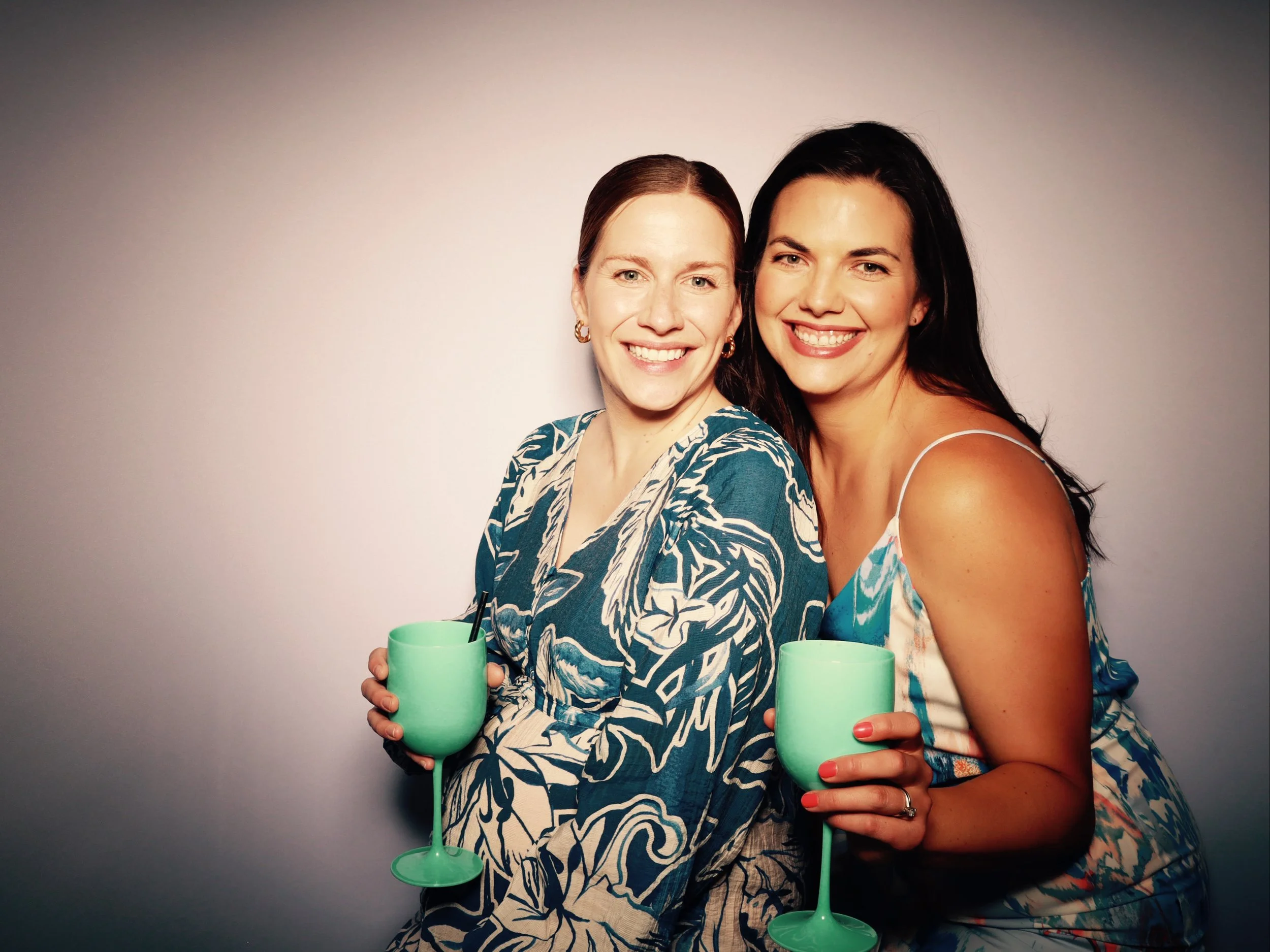 Two women smiling and holding green wine glasses against a plain background.