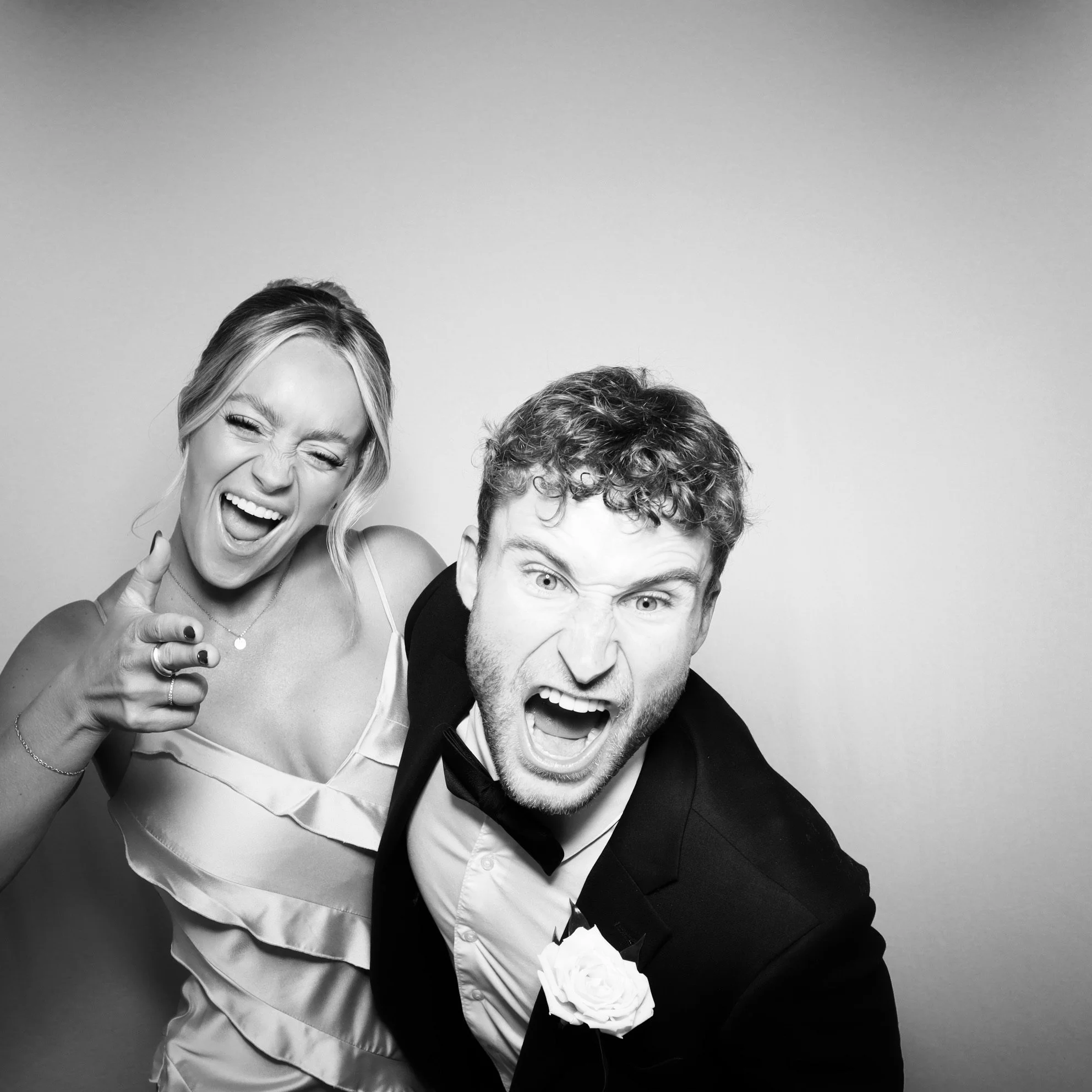 A black-and-white photo of a woman and a man with expressive faces, both pointing at the camera. The woman is smiling with her eyes closed, wearing a sleeveless dress, while the man is wearing a tuxedo with a flower lapel pin, and has his mouth open 