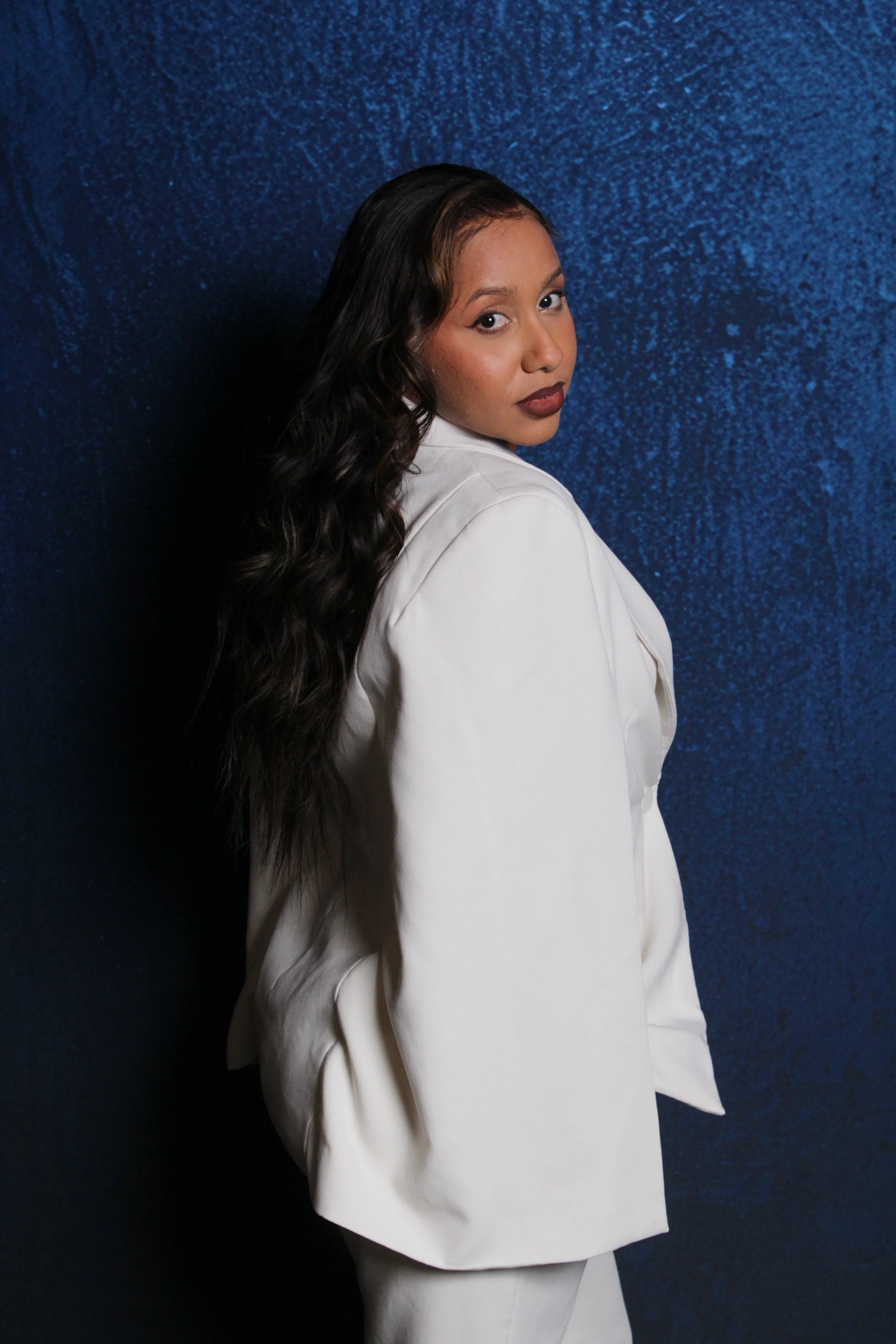 A woman with long dark wavy hair wearing a white blazer and pants poses against a dark blue textured background.